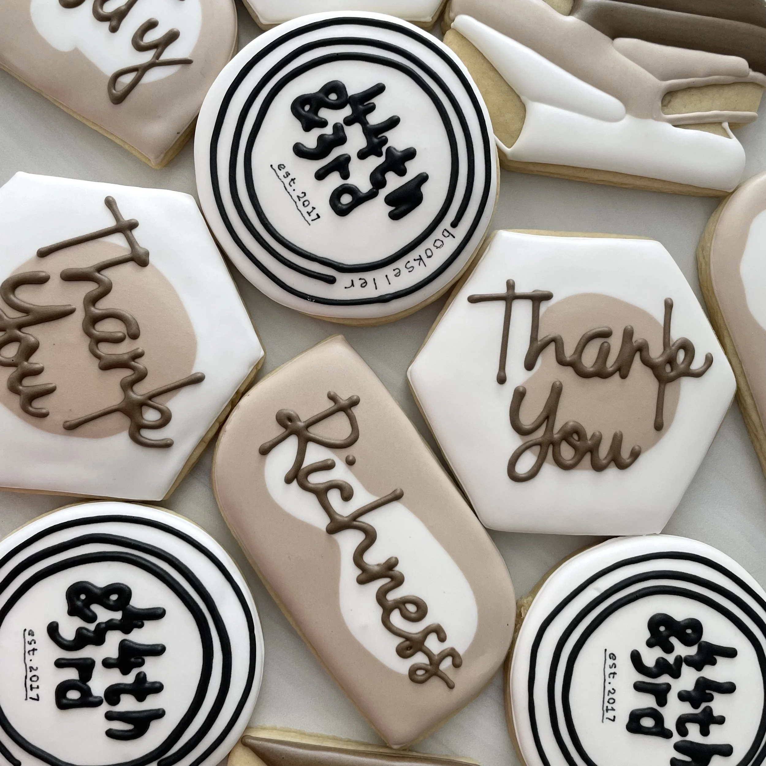 A variety of cookies decorated with white, black, and beige icing, featuring handwritten messages and patterns for a celebratory occasion.