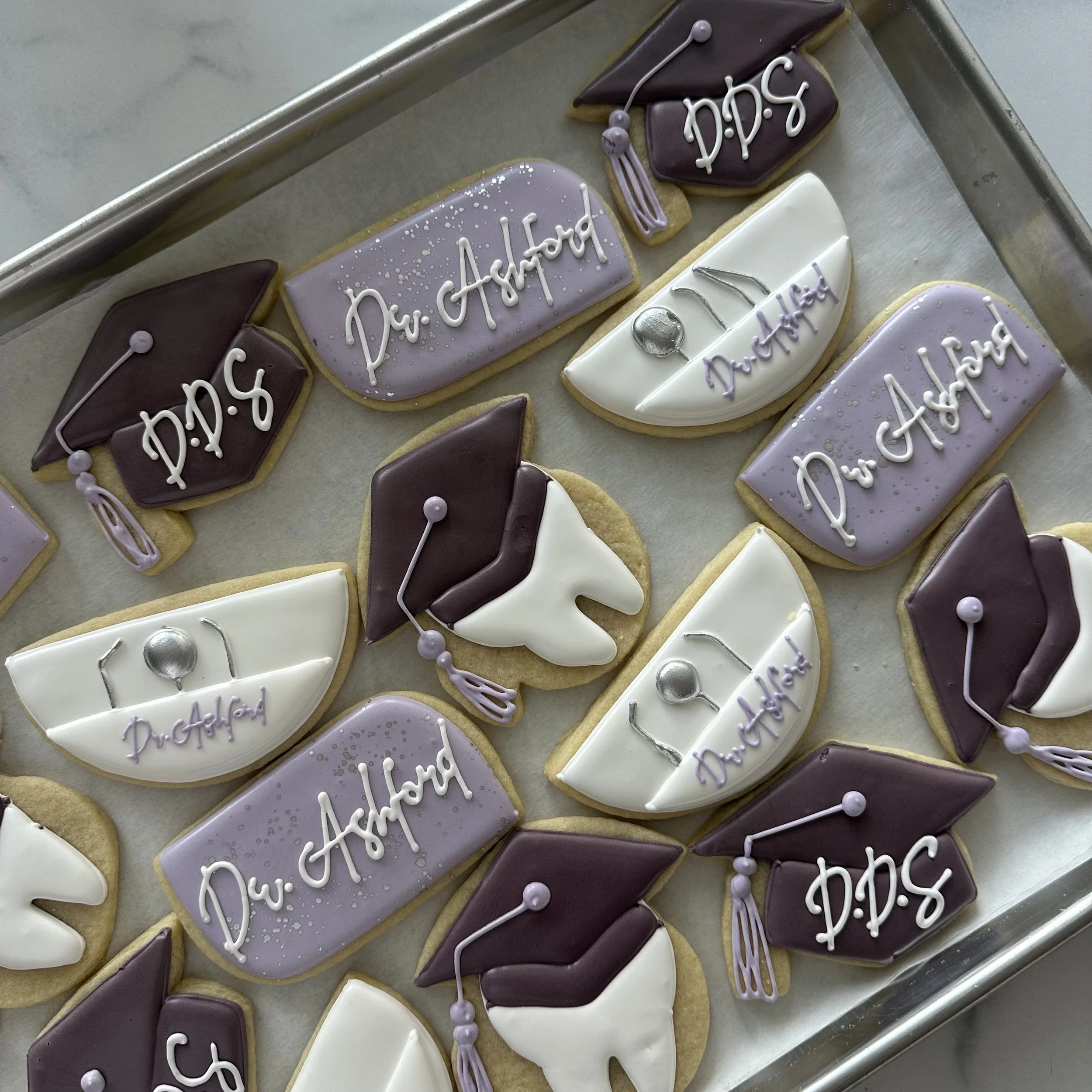 Decorated sugar cookies with graduation caps, diplomas, and the phrase 'Dr. Ashford' in purple and white icing, displayed on a baking sheet.