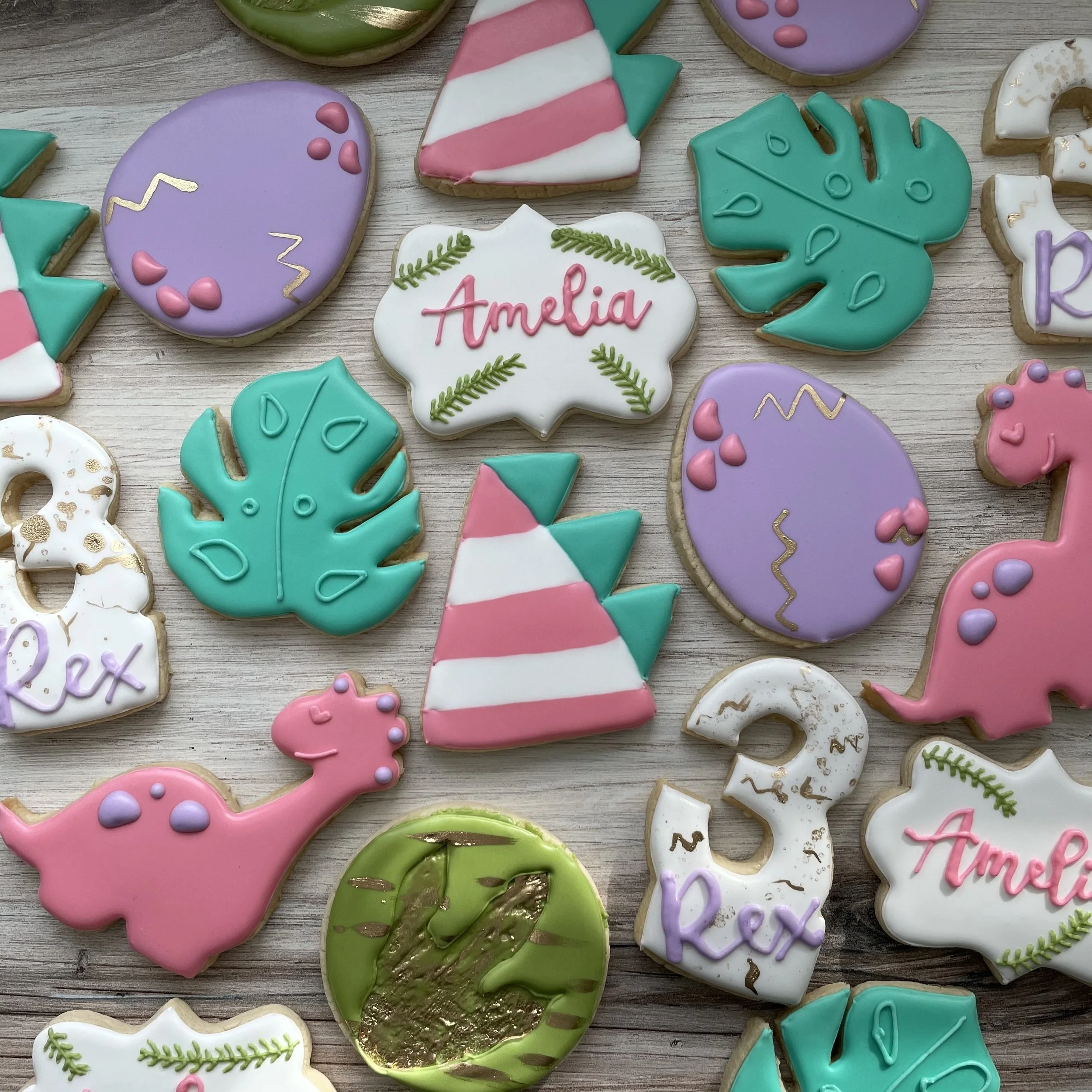 Assorted decorated sugar cookies on a wooden surface, including cookies with pink, purple, green, and white icing in various shapes such as leaves, hearts, and animal shapes, some with lettering and gold accents. One cookie has the name "Amelia" writ