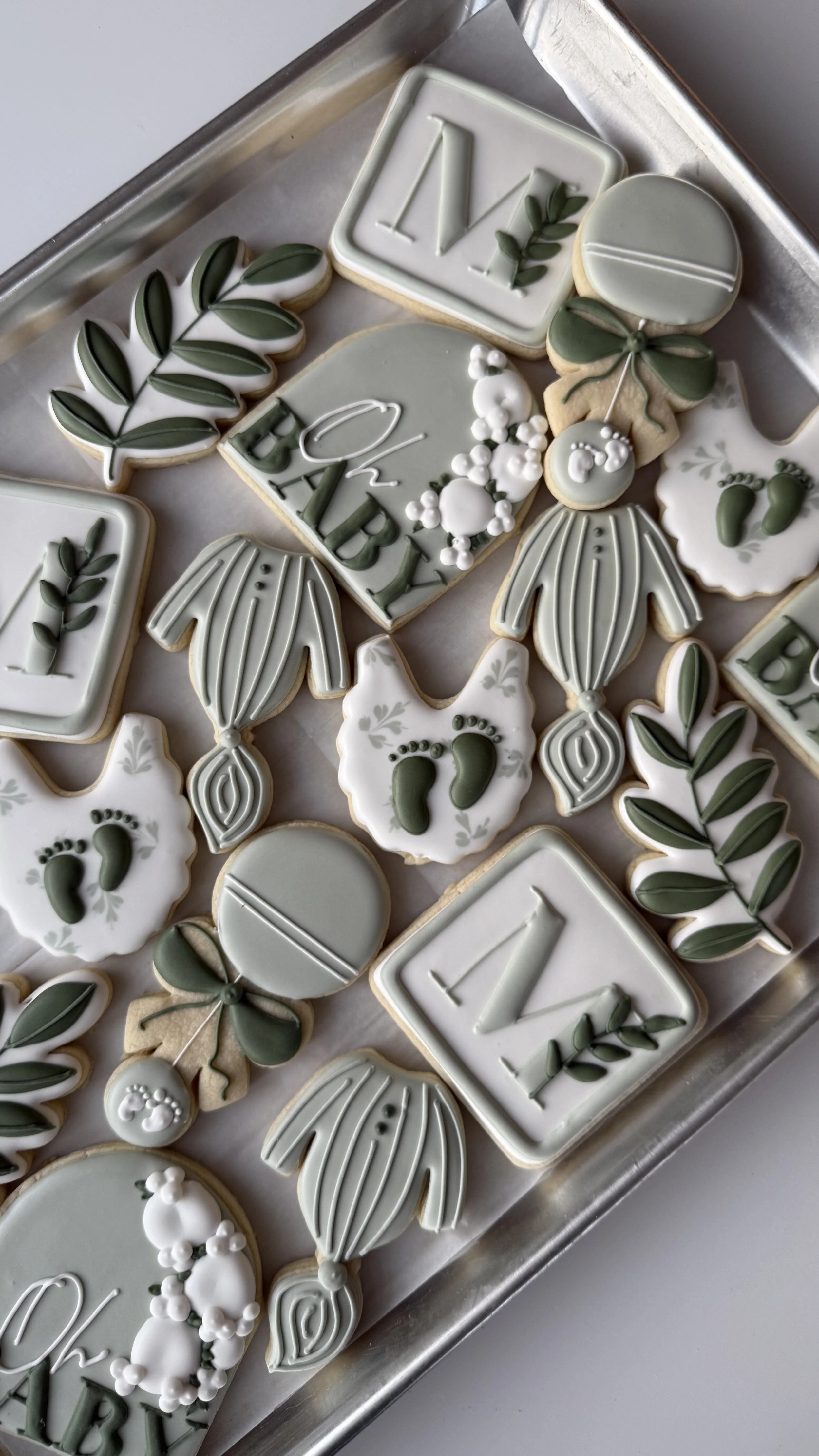 Assorted neutral-colored decorated cookies on a tray, including letters, leaves, baby footprints, and clothing designs.