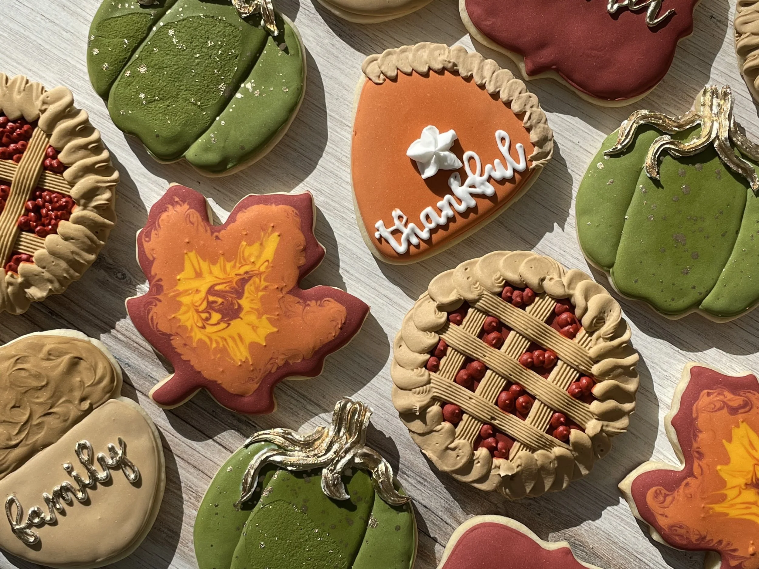 A variety of decorated Christmas cookies on a light wooden surface, including a green mitten-shaped cookie, an orange heart-shaped cookie with white and orange letters that spell 'thankful,' and other holiday-themed shapes with colorful icing and top
