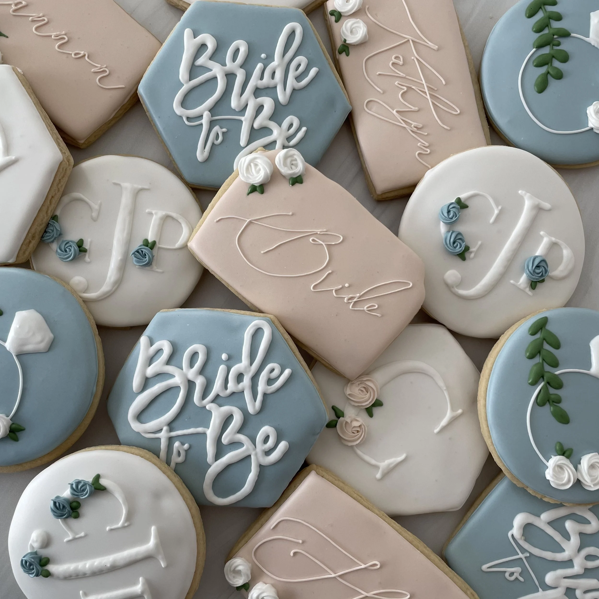 Decorated cookies with pastel blue, pink, white, and beige icing, featuring words like 'hello' and 'love,' with floral and swirl accents, arranged closely together.