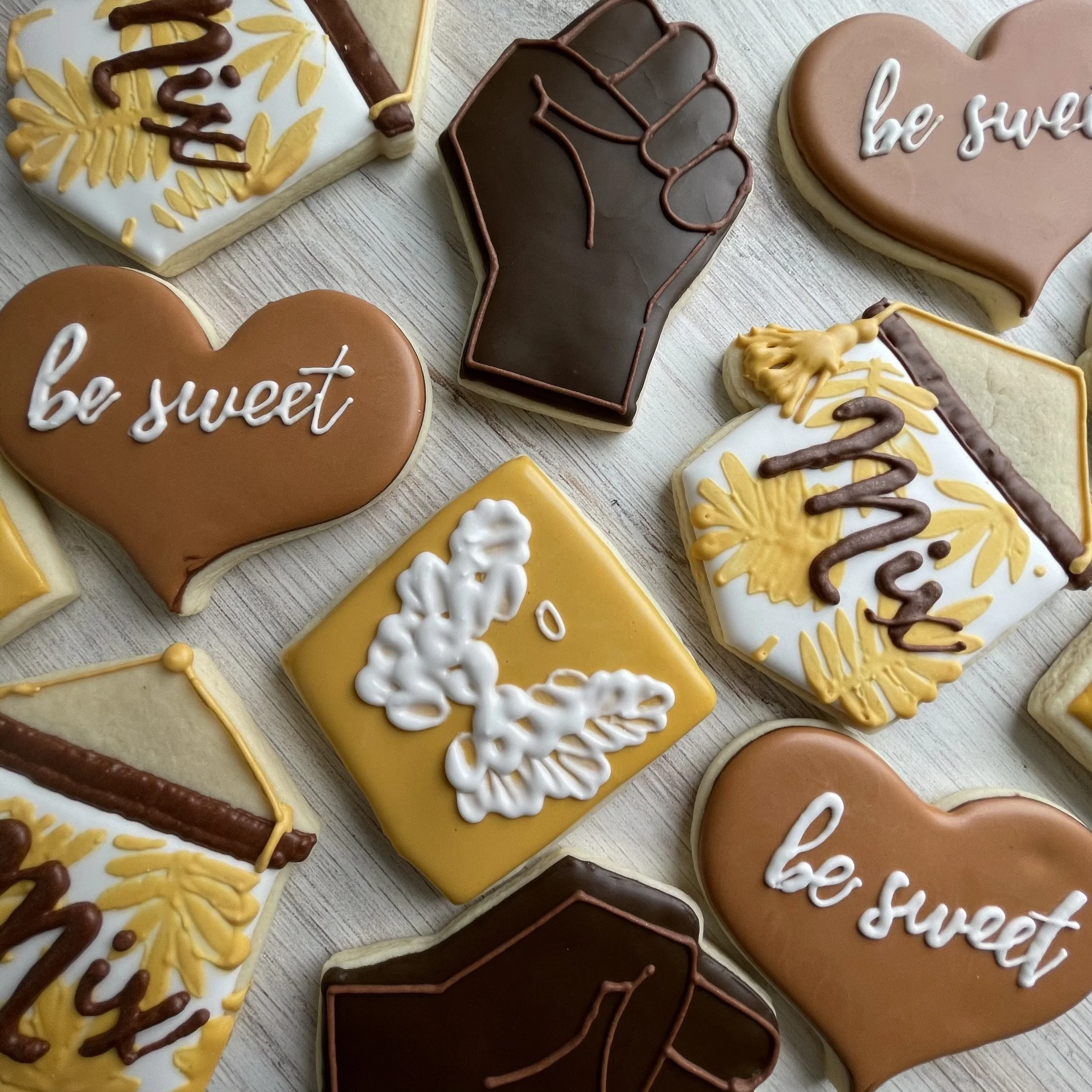 Decorated sugar cookies with phrases 'be sweet' and 'be you,' featuring hearts, flowers, and leaf patterns, decorated with icing in brown, yellow, white, and pink colors.