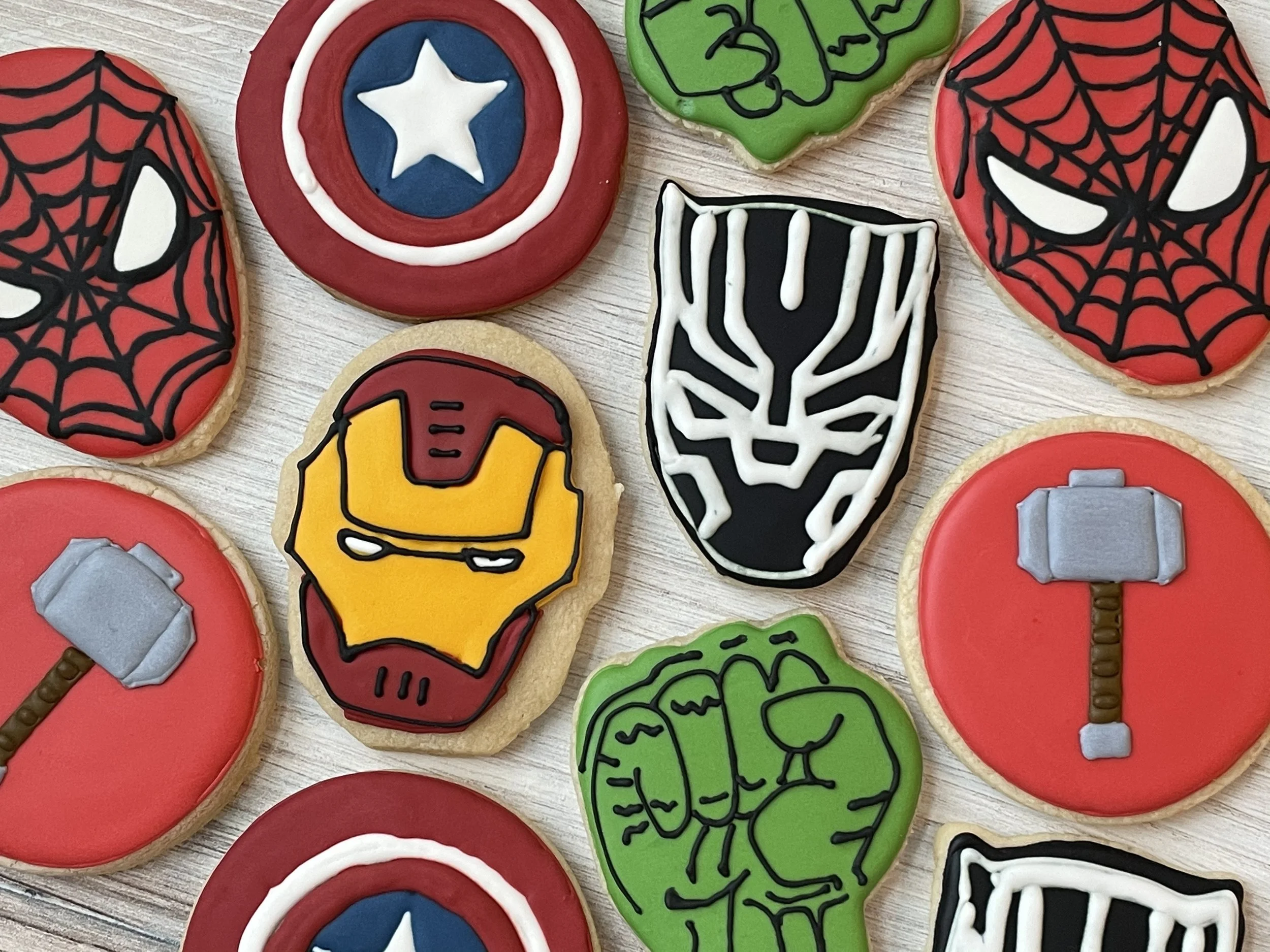 Colorful superhero-themed cookies decorated with designs inspired by Marvel characters such as Spider-Man, Iron Man, Captain America, and Venom, arranged on a light wooden surface.
