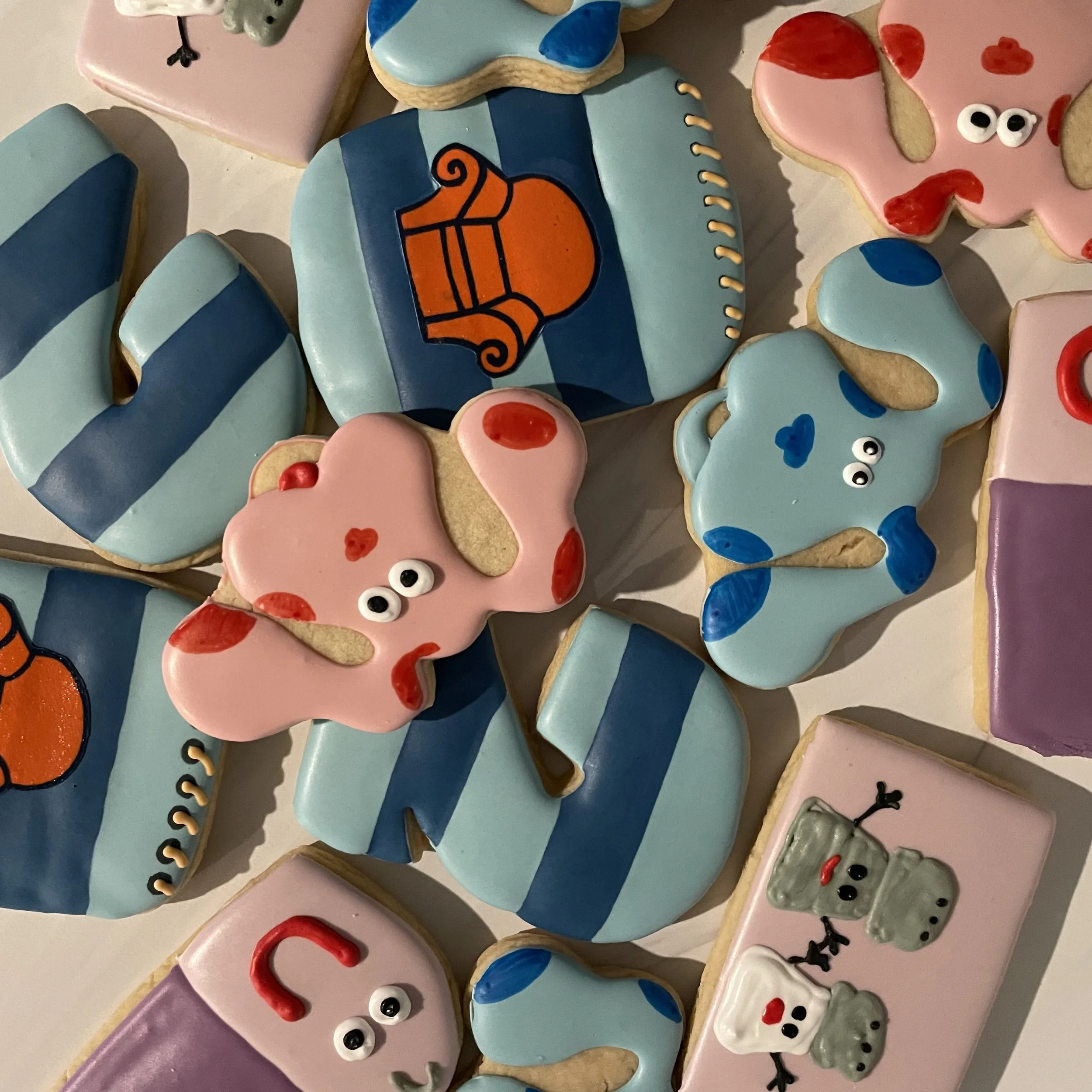 Assorted decorated sugar cookies with pink, blue, purple, and white icing, featuring designs such as roosters, stripes, and cartoonish eyes, arranged on a flat surface.