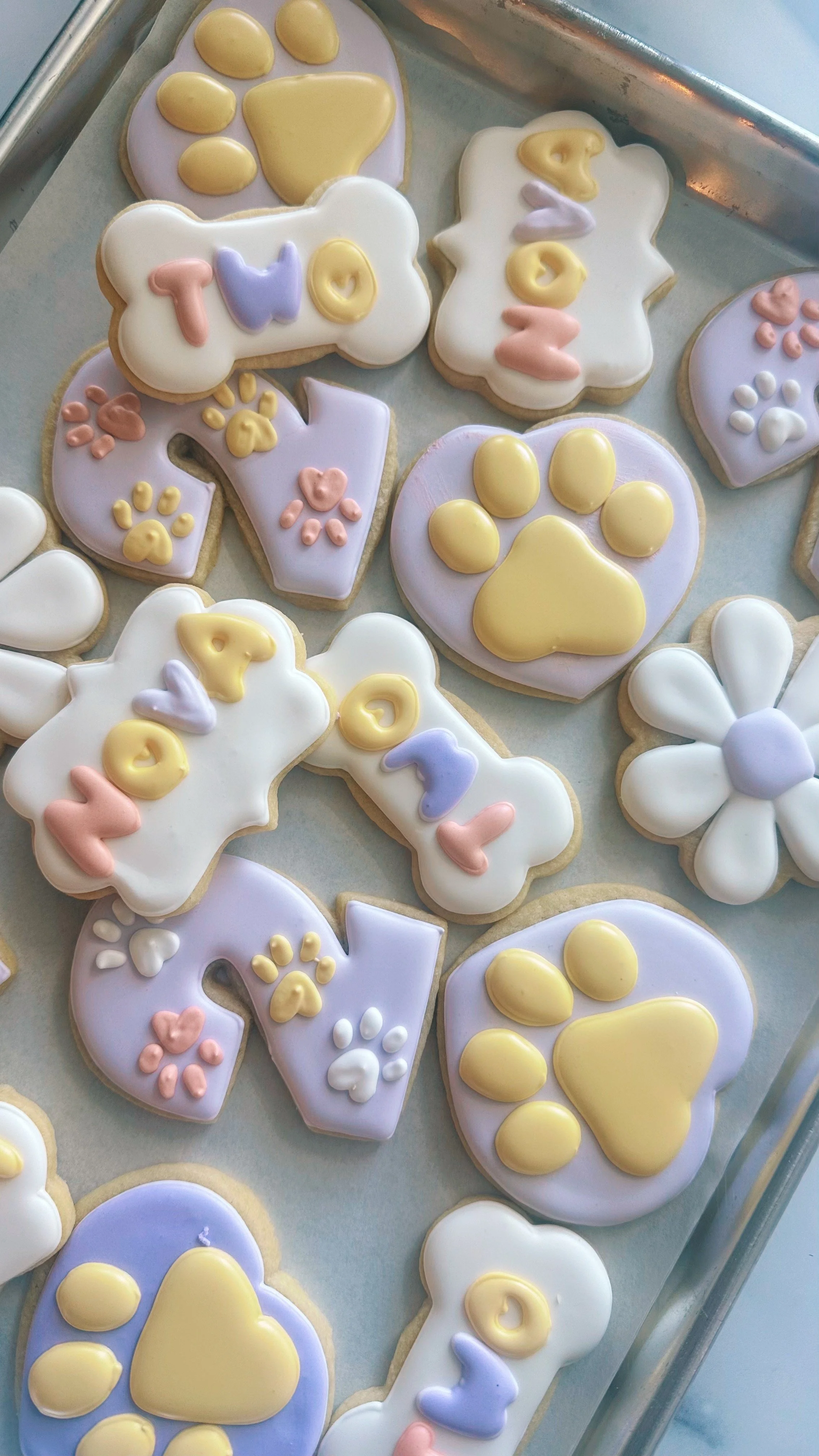 Cookies decorated with pastel-colored icing in the shapes of paws and flowers, with the words 'TOO' and paw prints.