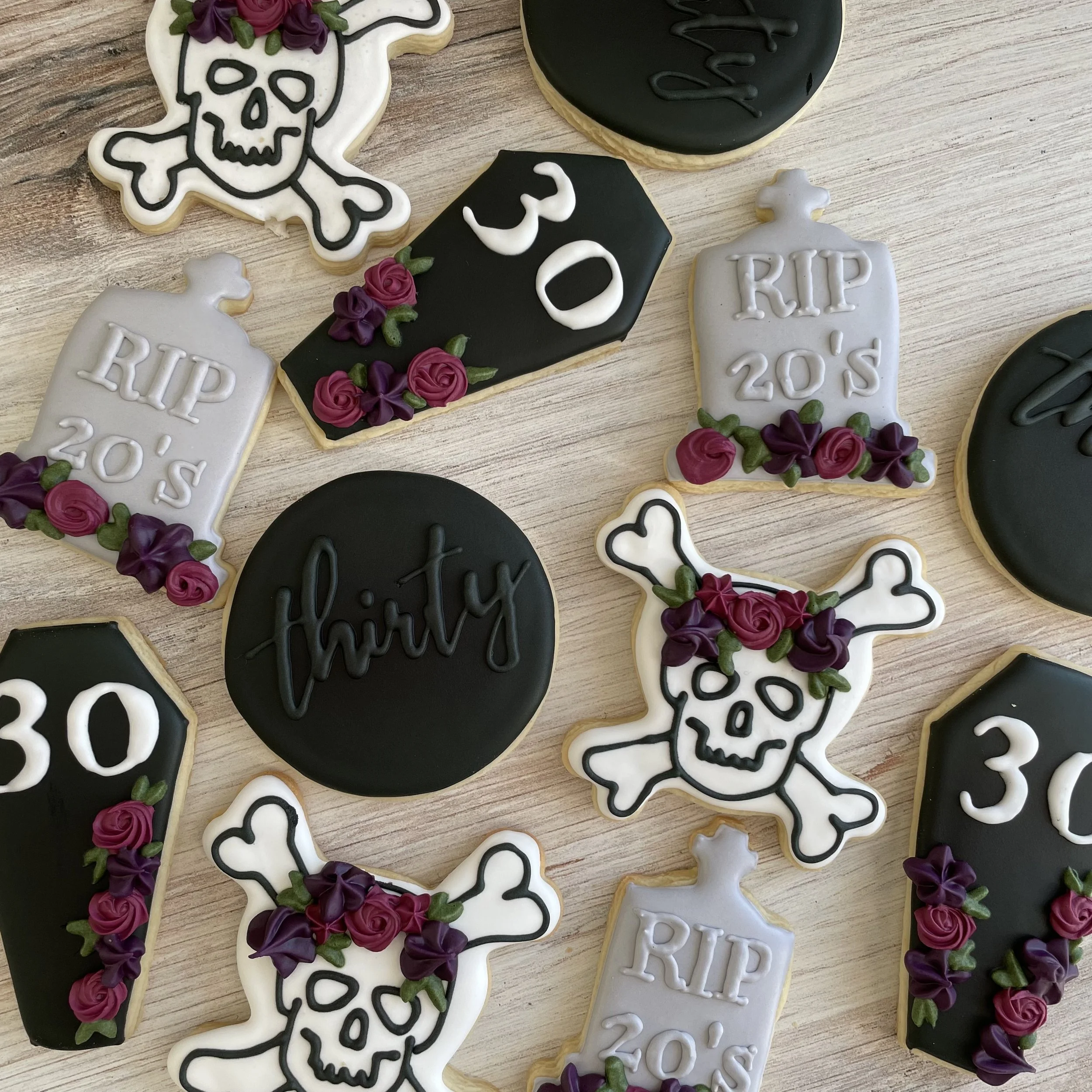 Decorative cookies with Halloween themes, including skulls, coffins, and gravestones with flowers in purple and green, on a wooden surface.