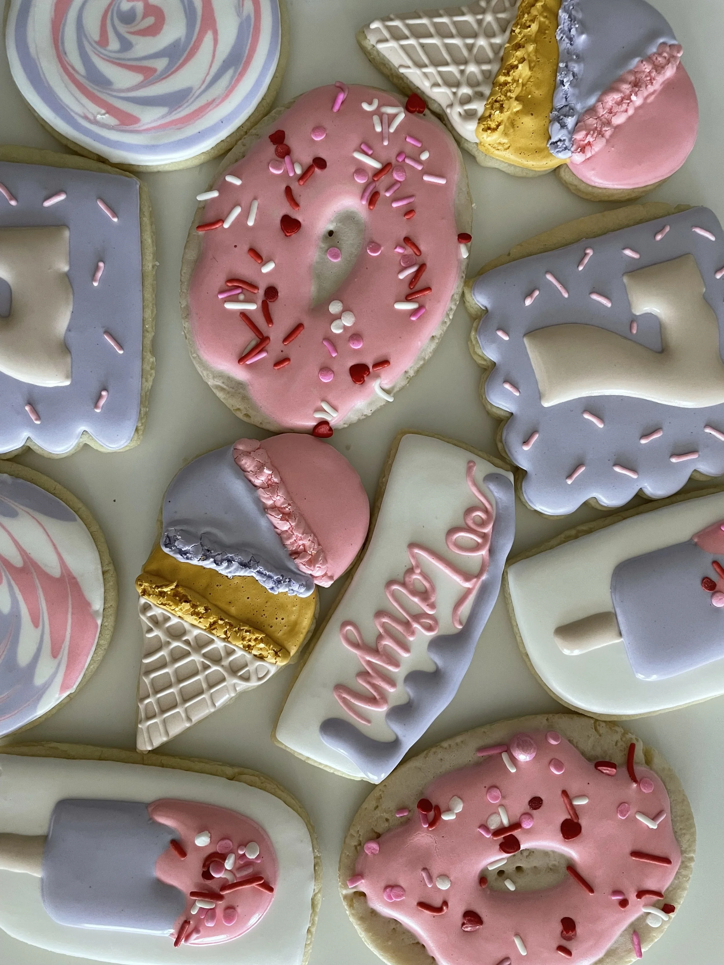 Collection of decorated sugar cookies in various shapes including a heart, a letter, and a rainbow, decorated with pink, gray, white, yellow, and purple icing and sprinkles.