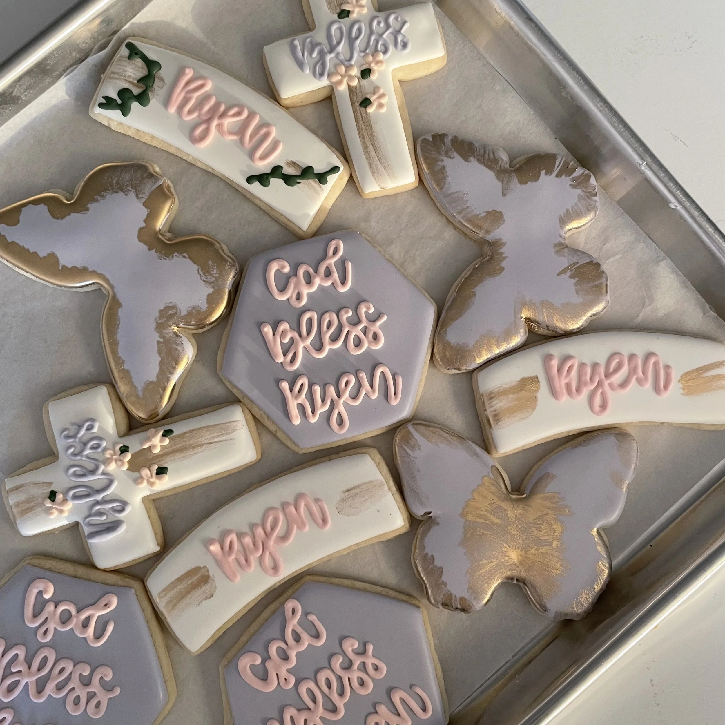 Assorted decorated cookies on a baking sheet. Some cookies have white, pink, and purple icing, with words like 'thank you' and decorative floral and foliage designs.