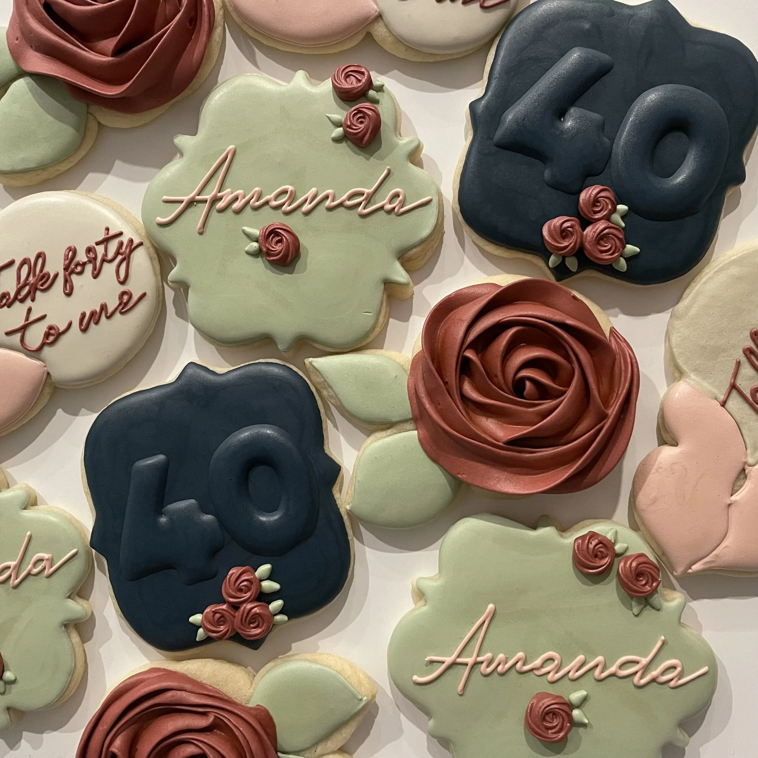 Assorted cookies decorated with pastel-colored icing, including cookies with the words "Amanda" and "You" and decorated with roses and cursive writing.