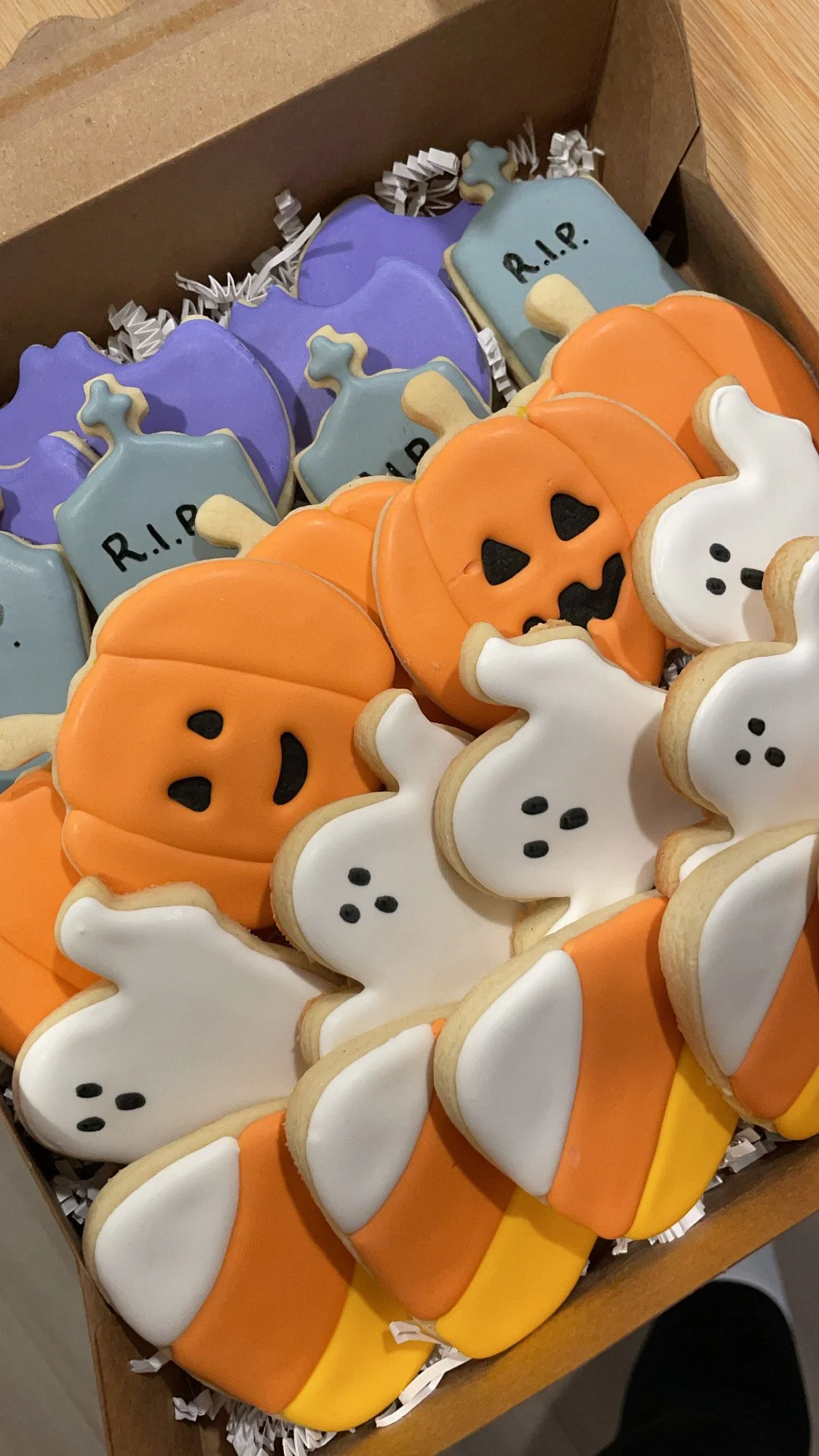 Halloween-themed decorated cookies in the shape of pumpkins, ghosts, and tombstones decorated with colored icing and black details, arranged in a box.