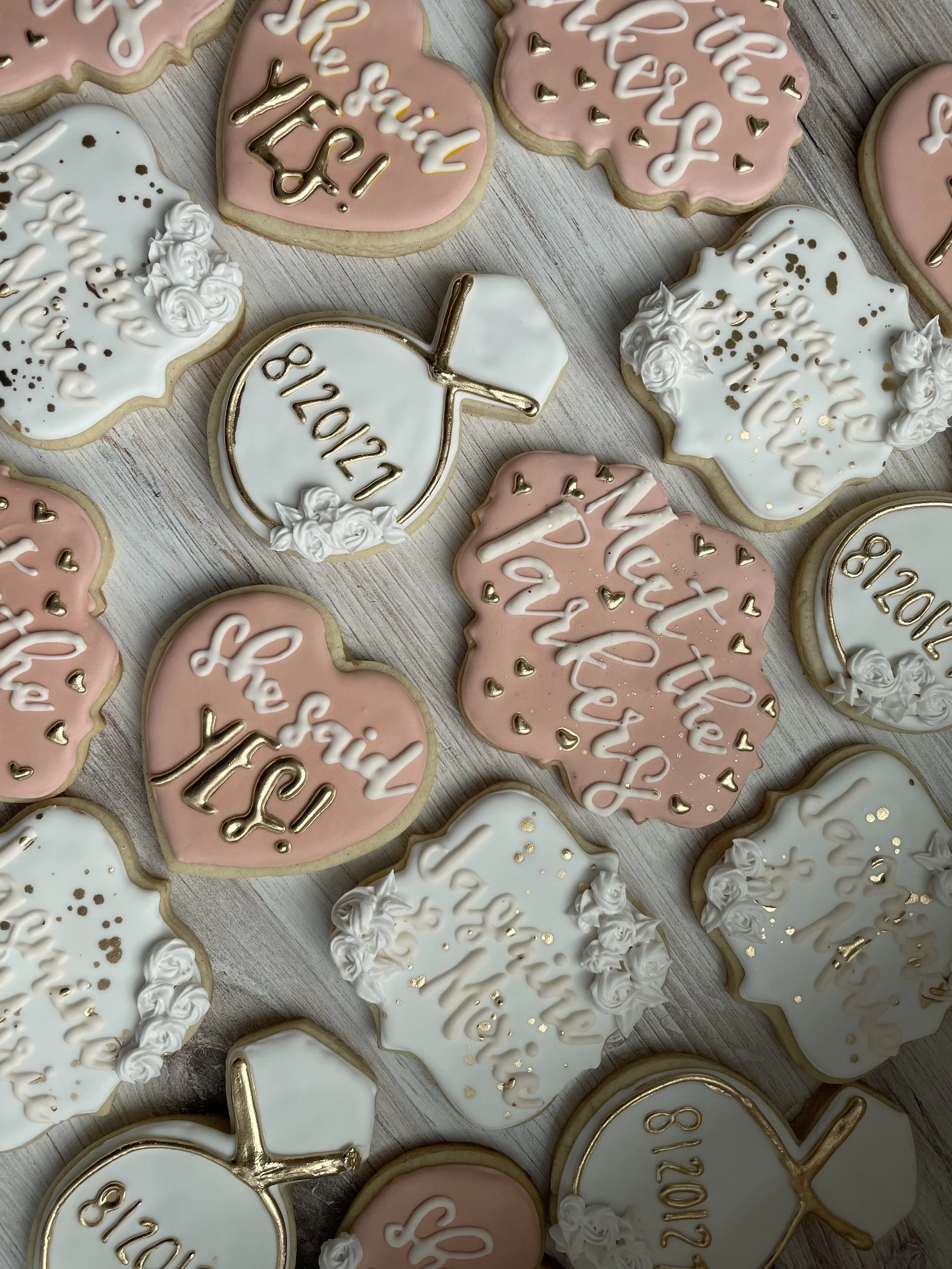 Assorted decorated cookies with pink and white icing, gold accents, and messages such as "Thank You!" and "8/20/2021," along with cookies shaped like apples and hearts.