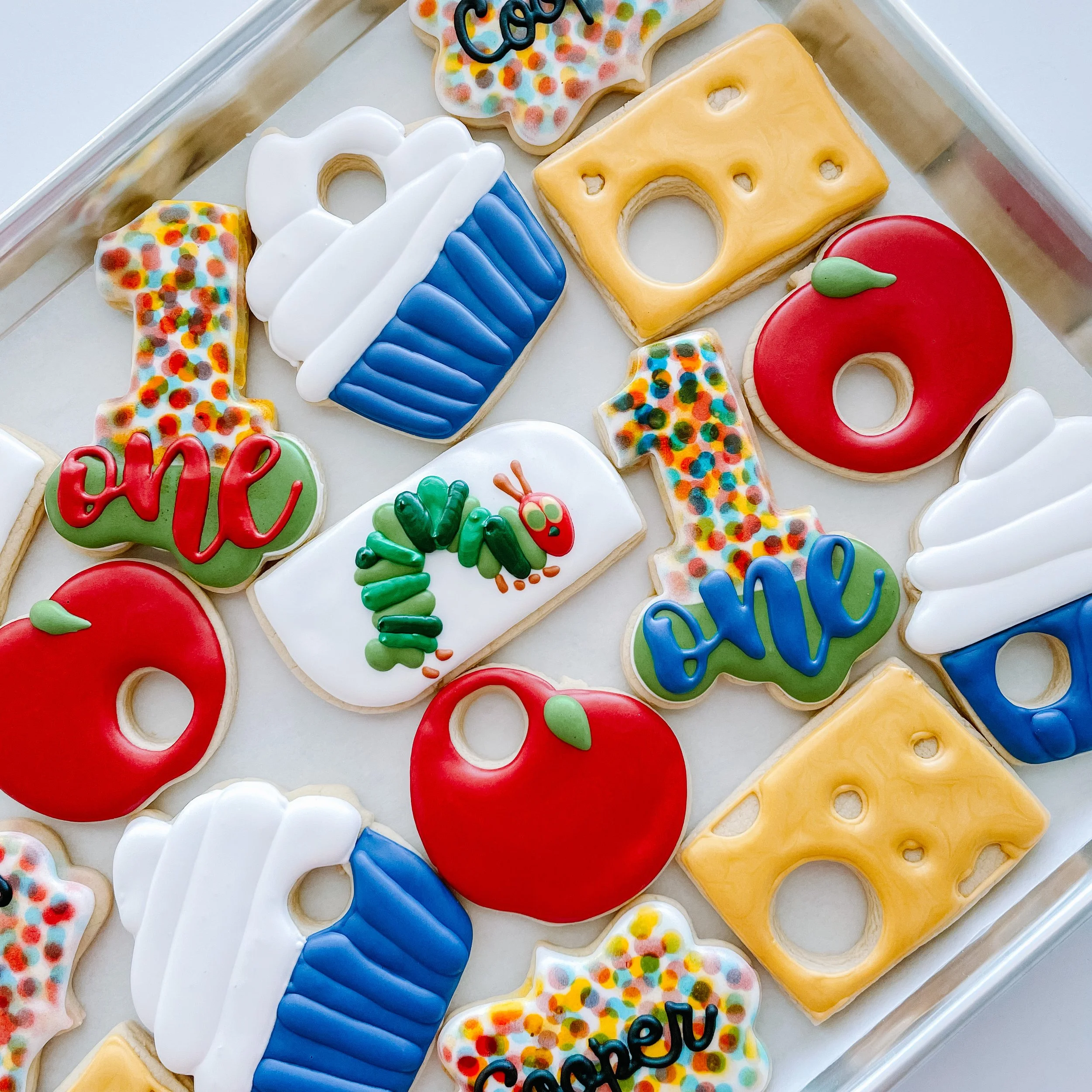 Colorful birthday cookies on a tray, including cupcakes, a yellow cheese-shaped cookie, a red apple cookie, and other decorated treats with sprinkles and lettering.