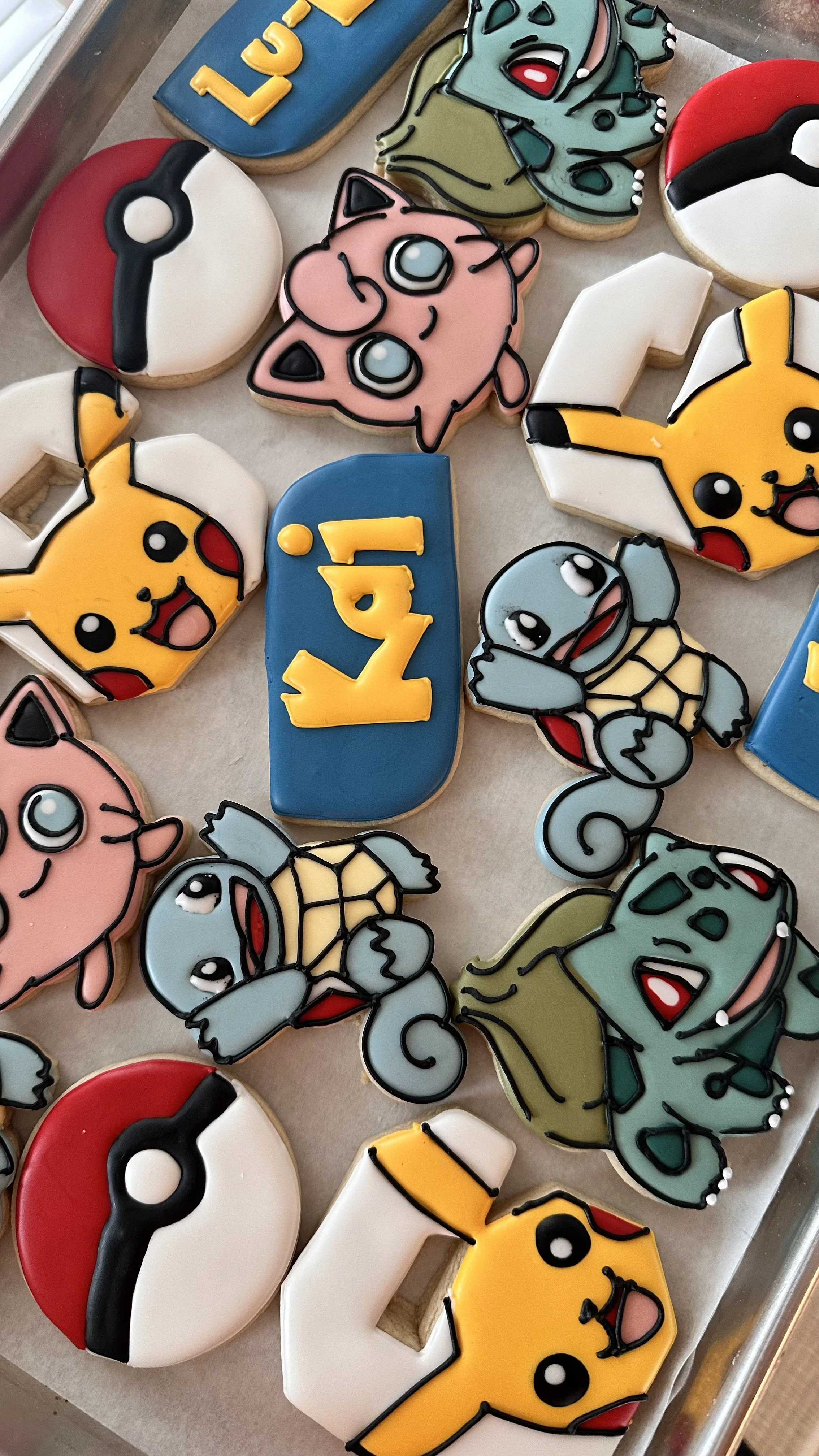 Various decorated cookies themed around Pokémon, including Pikachu, Eevee, Squirtle, Bulbasaur, Jigglypuff, and Pokéball, arranged on a tray.