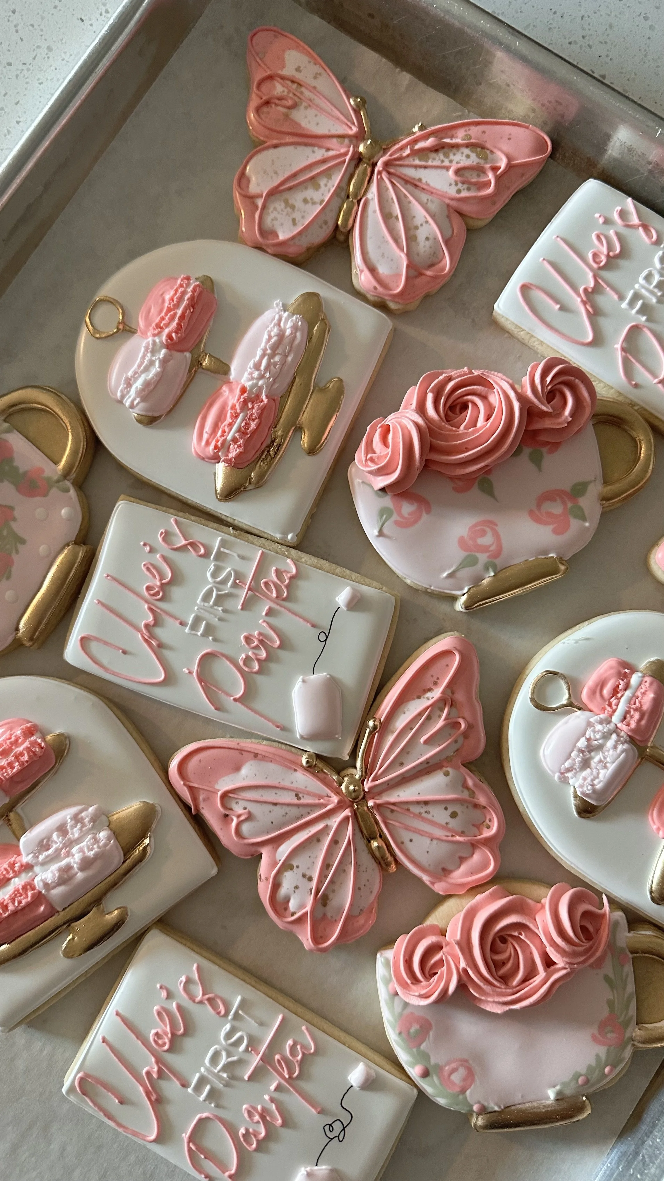 Decorative cookies in various shapes including butterflies, teacups, and airplanes with pink, white, and gold icing, arranged on a tray.