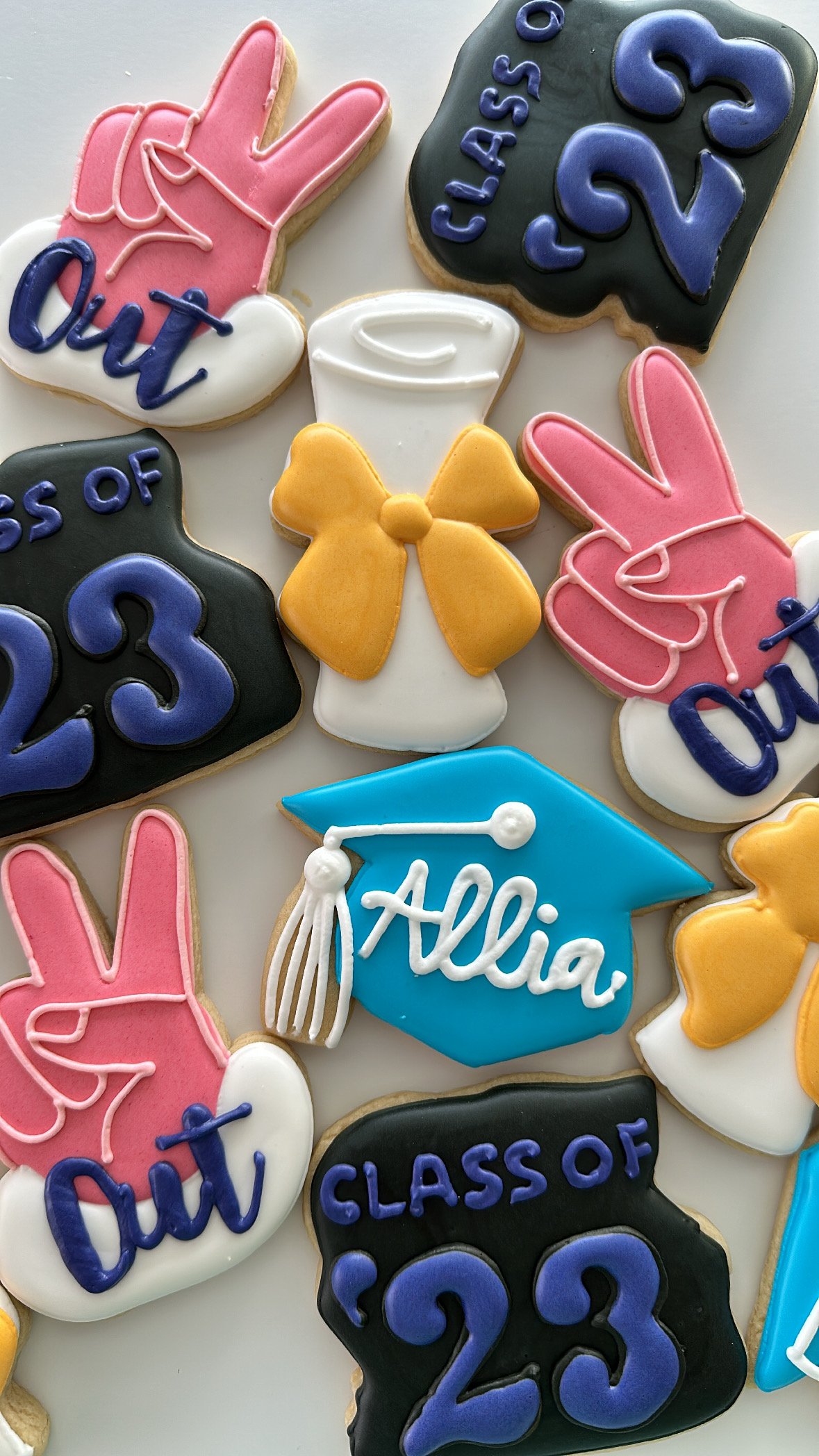 Decorated cookies themed for graduation with messages like "Class of 2023," featuring traditional elements like sashes, a mortarboard with "2023," and symbols such as peace signs and bows.