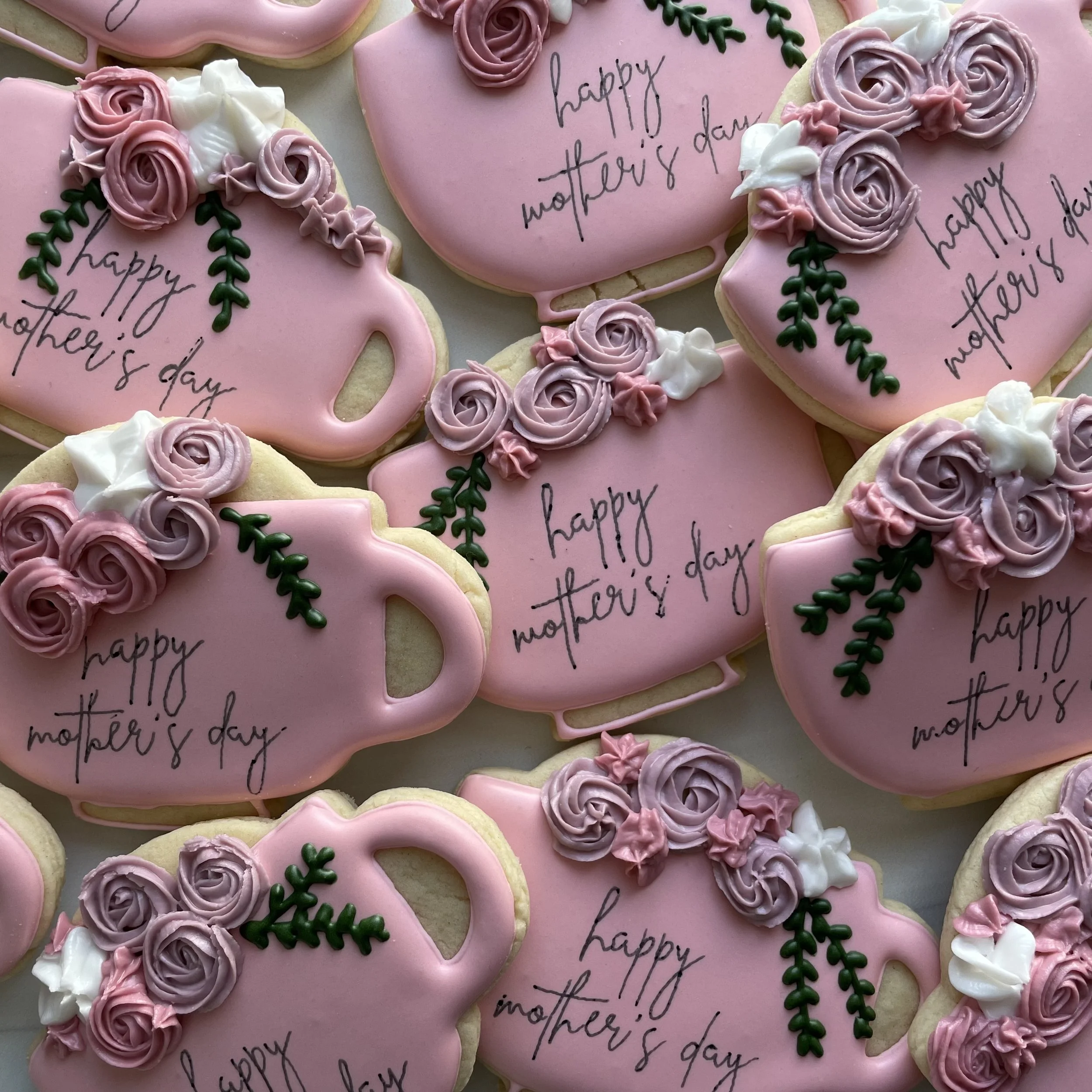 Decorated heart-shaped cookies with pink, white, and purple frosting, floral designs, and handwritten messages reading "happy mother's day" on light pink background.