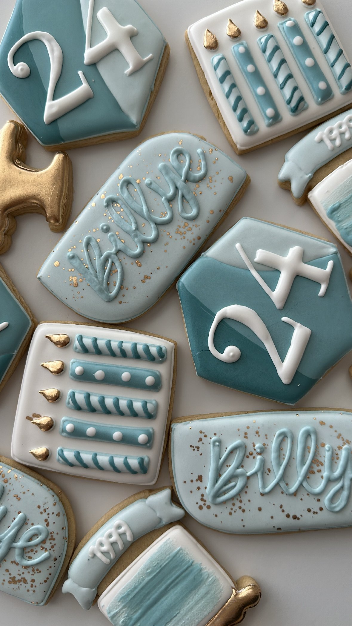 Cake cookies decorated with blue, white, and gold icing, including designs like stars, text, and patterns, celebrating a baby shower with a blue and gold color theme.