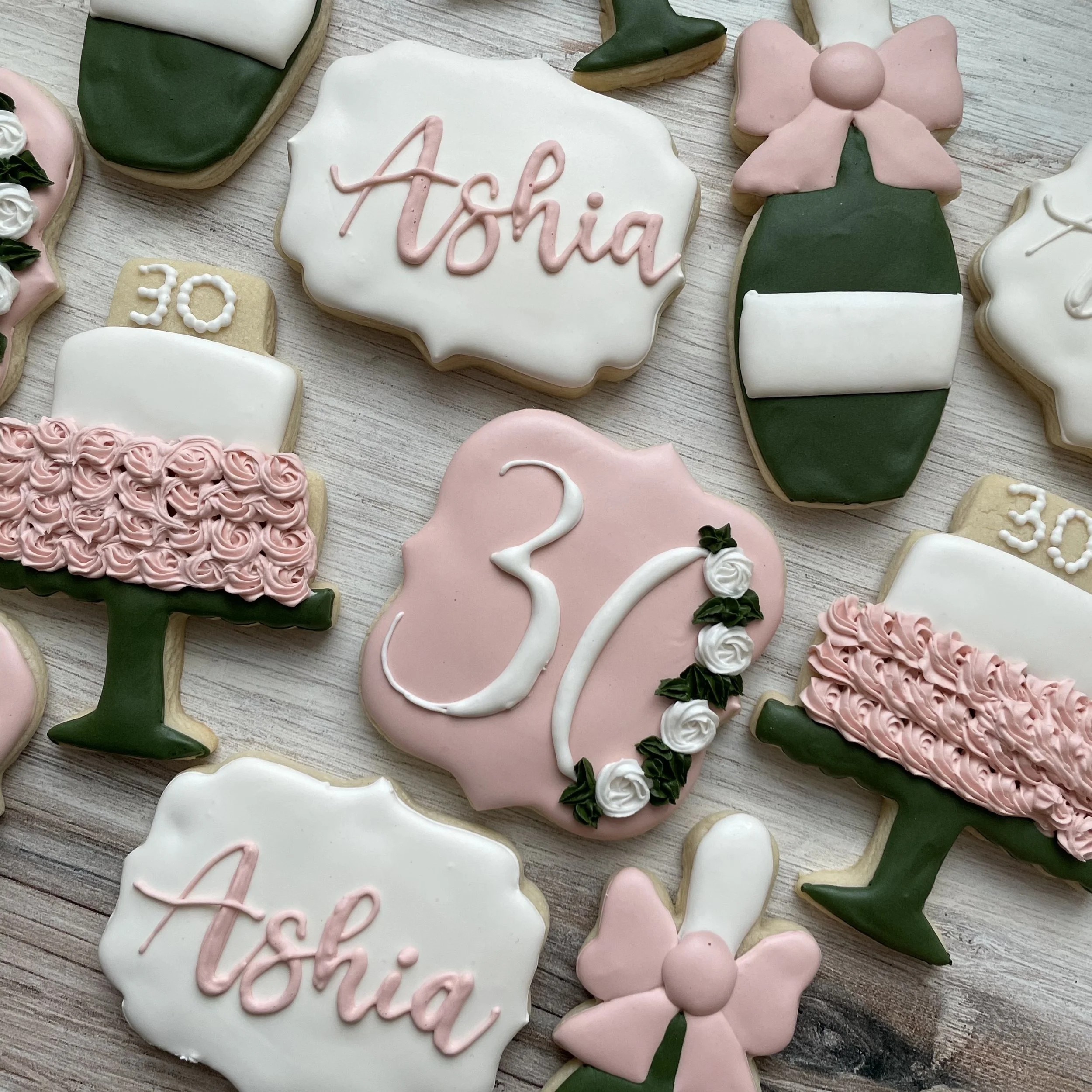 Decorative cookies for a birthday celebration, including cookies with 'Alice' written in pink icing, floral designs, and pink bow shapes on a wooden surface.