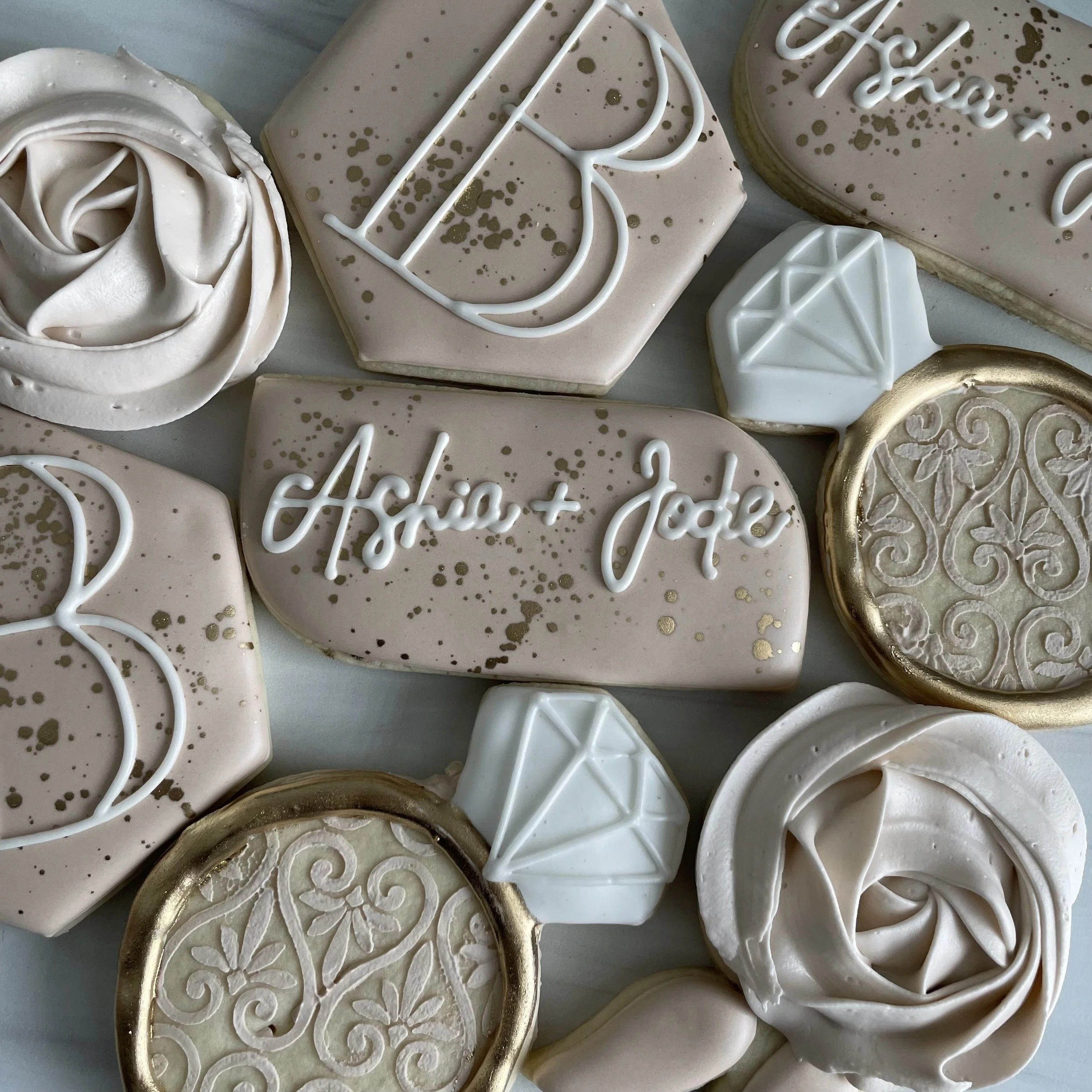 Assorted decorated cookies, including ones with white and gold icing, swirls, edible gold splashes, and handwritten messages.