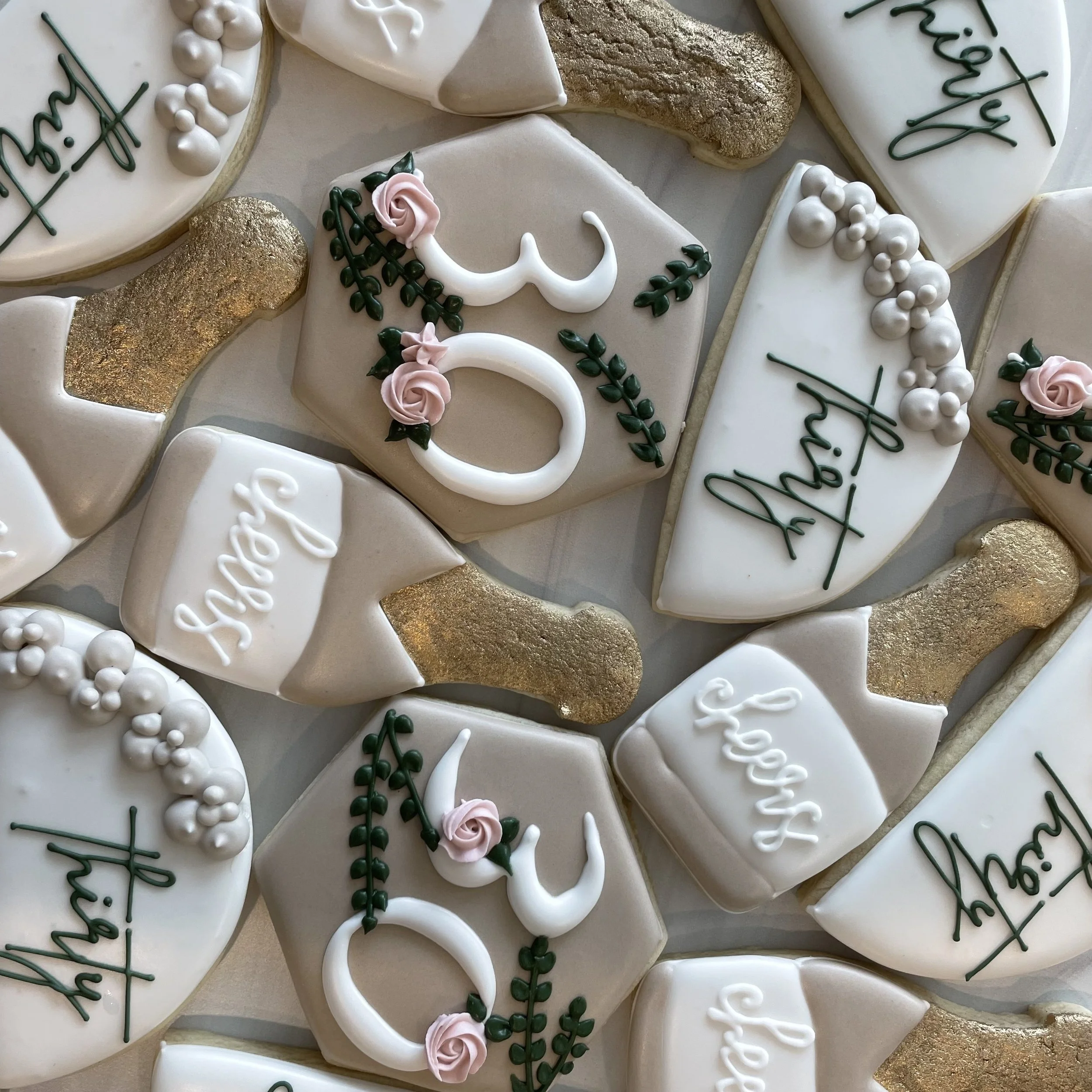 Close-up of decorated cookies with white, gold, and pink icing, featuring floral and message designs for a celebration.