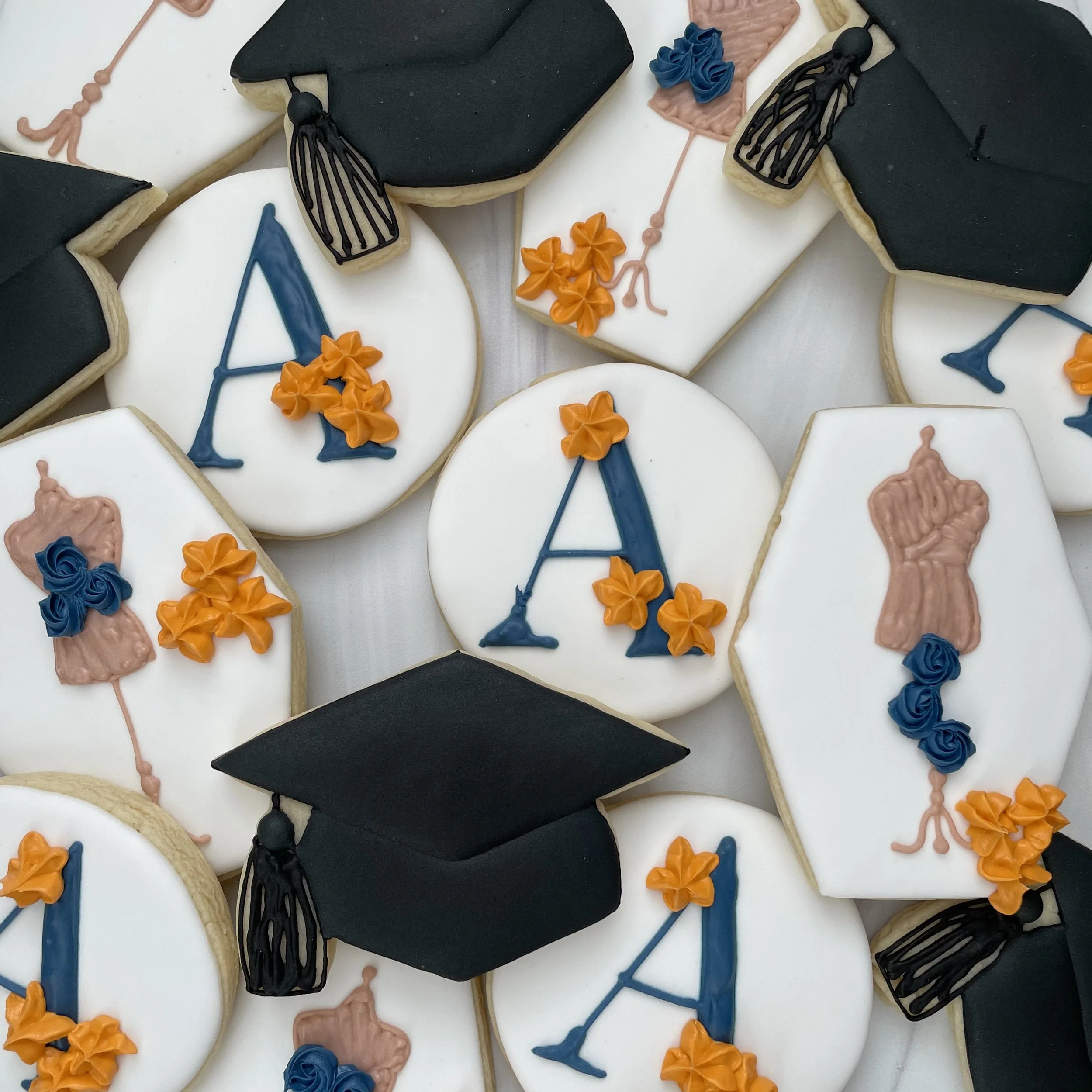 Cookies decorated with black, orange, blue, and pink icing, some featuring graduation cap designs and others with floral decorations, on a white surface.