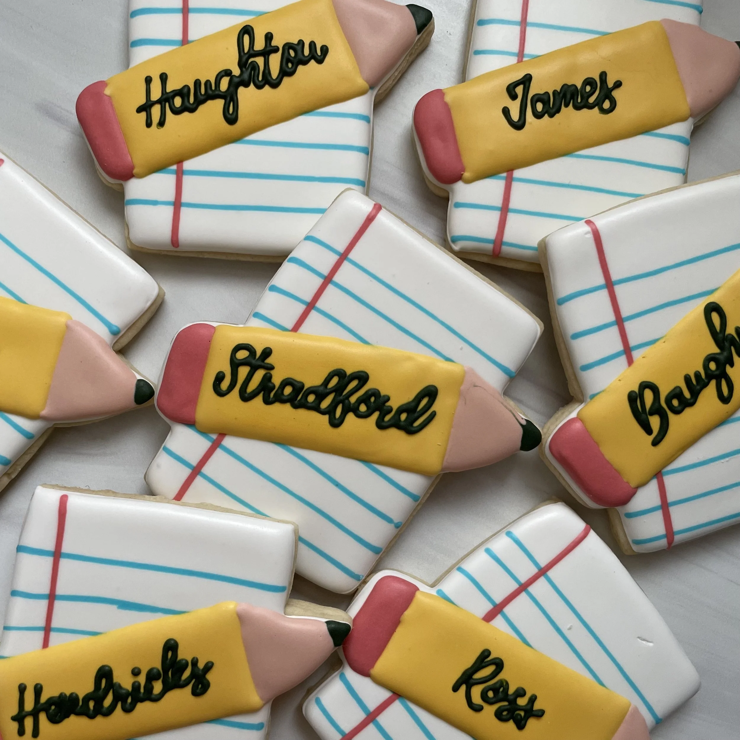Decorative cookies shaped like pencils with messages written on them, placed on a surface with other similar cookies.