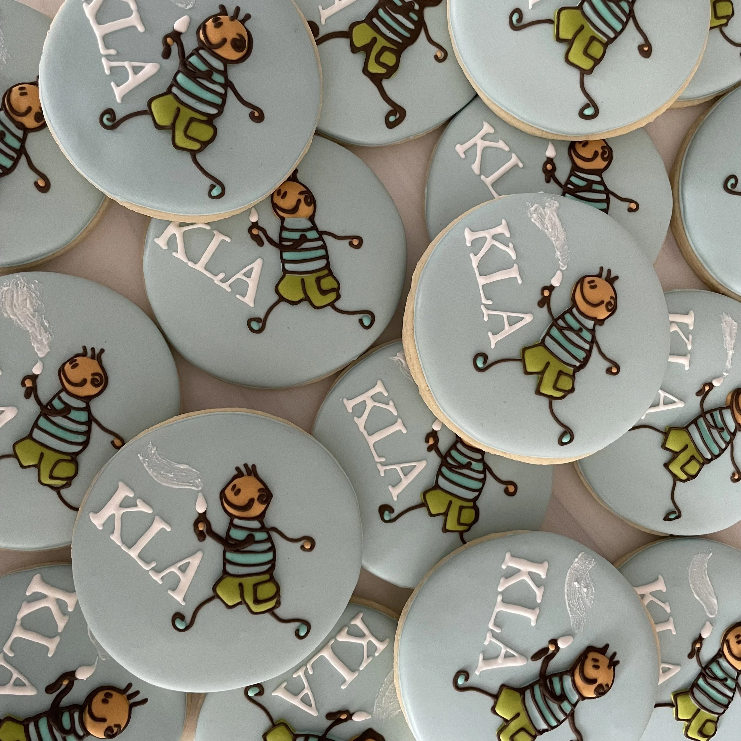 Cookies decorated with light blue icing featuring cartoon bees, white snowflakes, and the word 'KLAI' written in white.