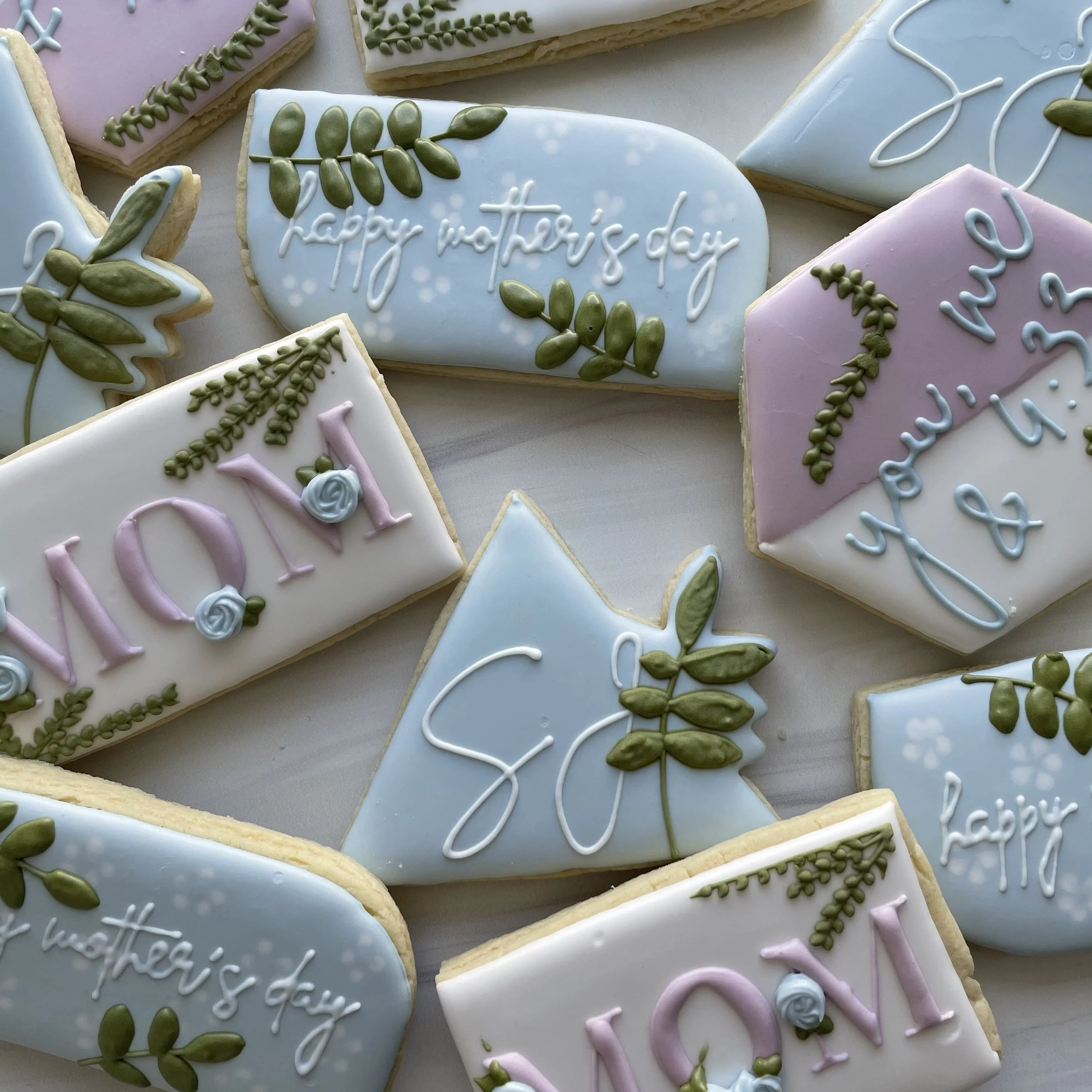 Assorted decorated cookies with pastel-colored icing including pink, blue, and white, featuring messages like 'happy mother’s day' and floral designs with green leaves, arranged on a neutral background.
