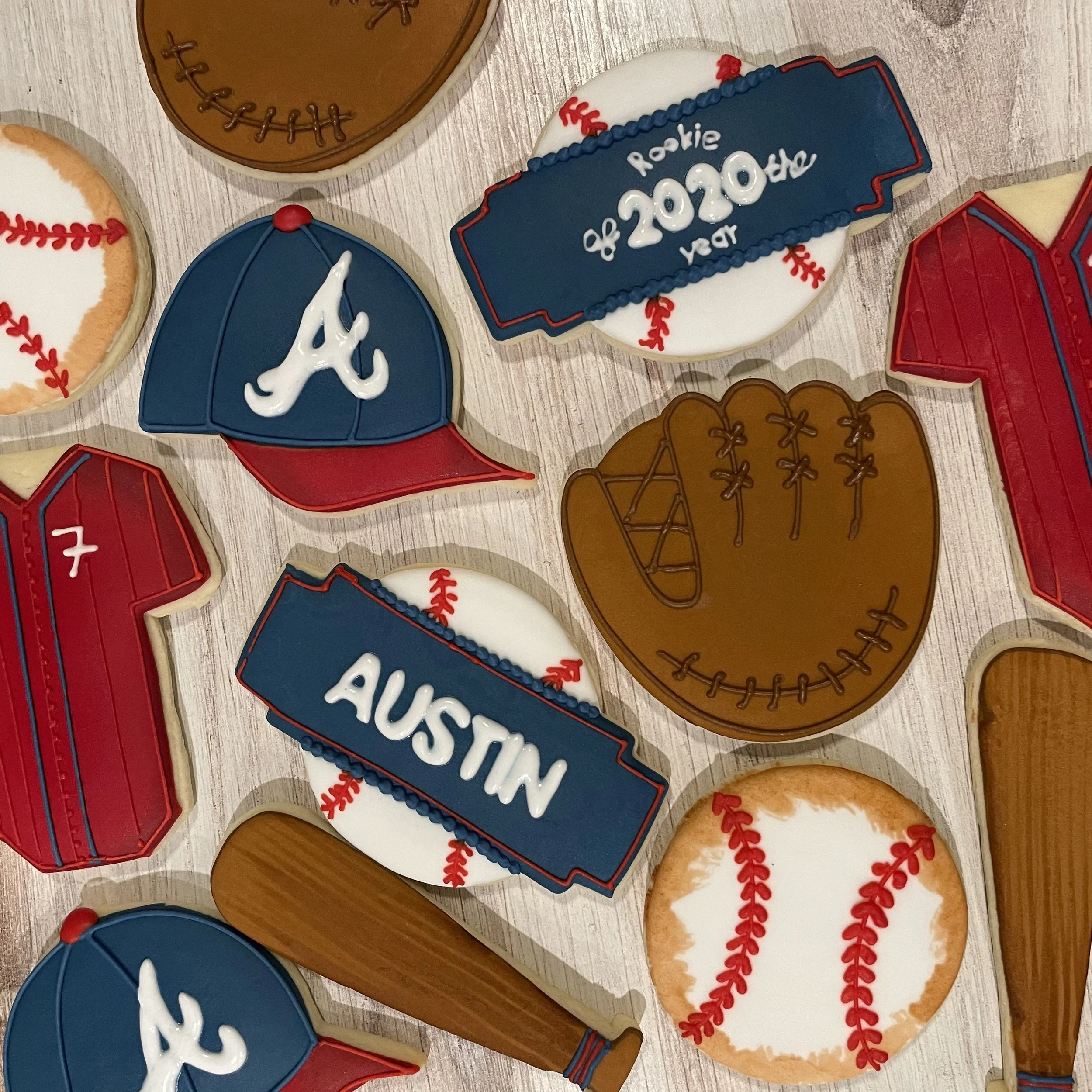 Assorted baseball-themed cookies decorated with baseball stitching, baseball gloves, and team logos in red, blue, white, and brown icing on a wooden surface.