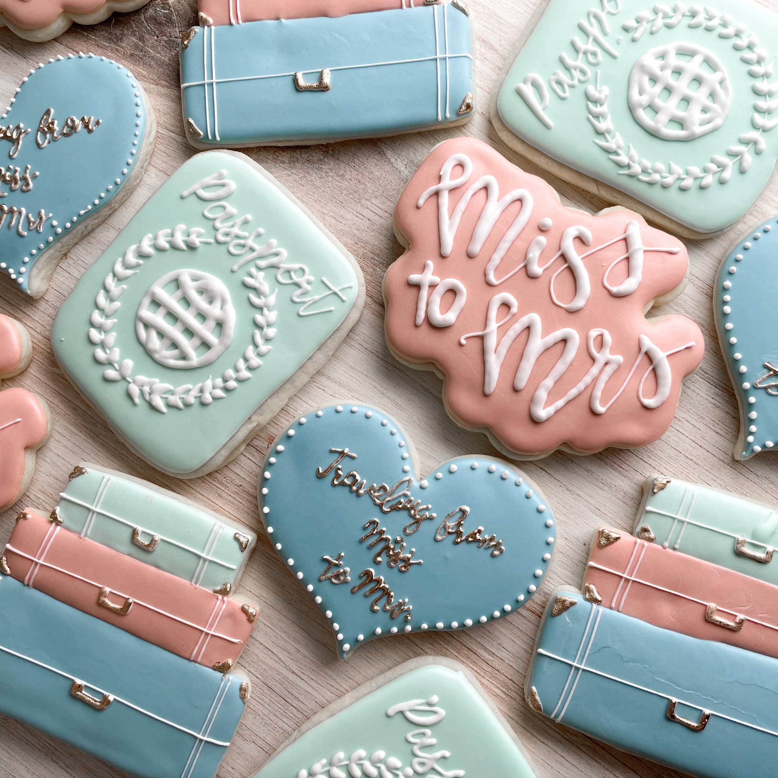Decorated cookies with pastel blue and pink icing, some shaped like hearts and rectangles, featuring white and silver writing and piping, on a light wooden surface.