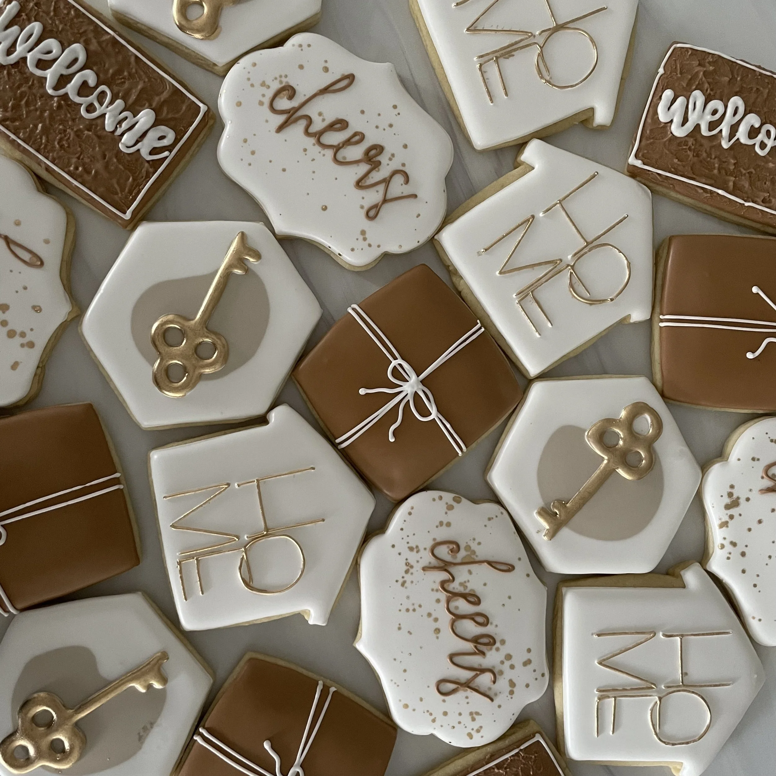 Assorted decorated cookies with messages and ornaments, including cookies that say 'welcome' and 'cheers,' some shaped like hearts and others with gold accents.