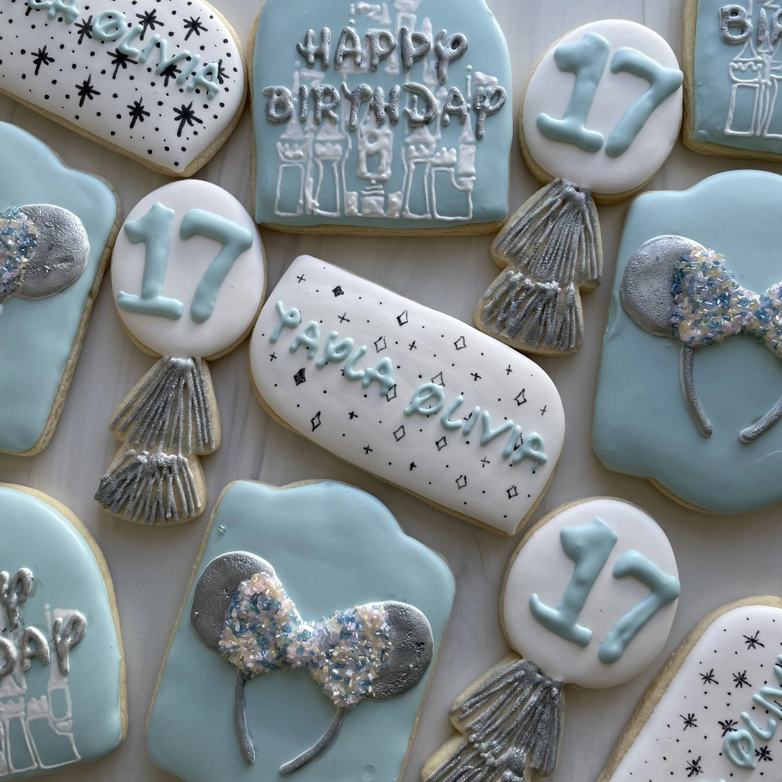 Cookies decorated with light blue, white, and silver icing. Some cookies have the number 12, others say 'happy birthday', and some are designed as balloons and presents, all for a birthday celebration.