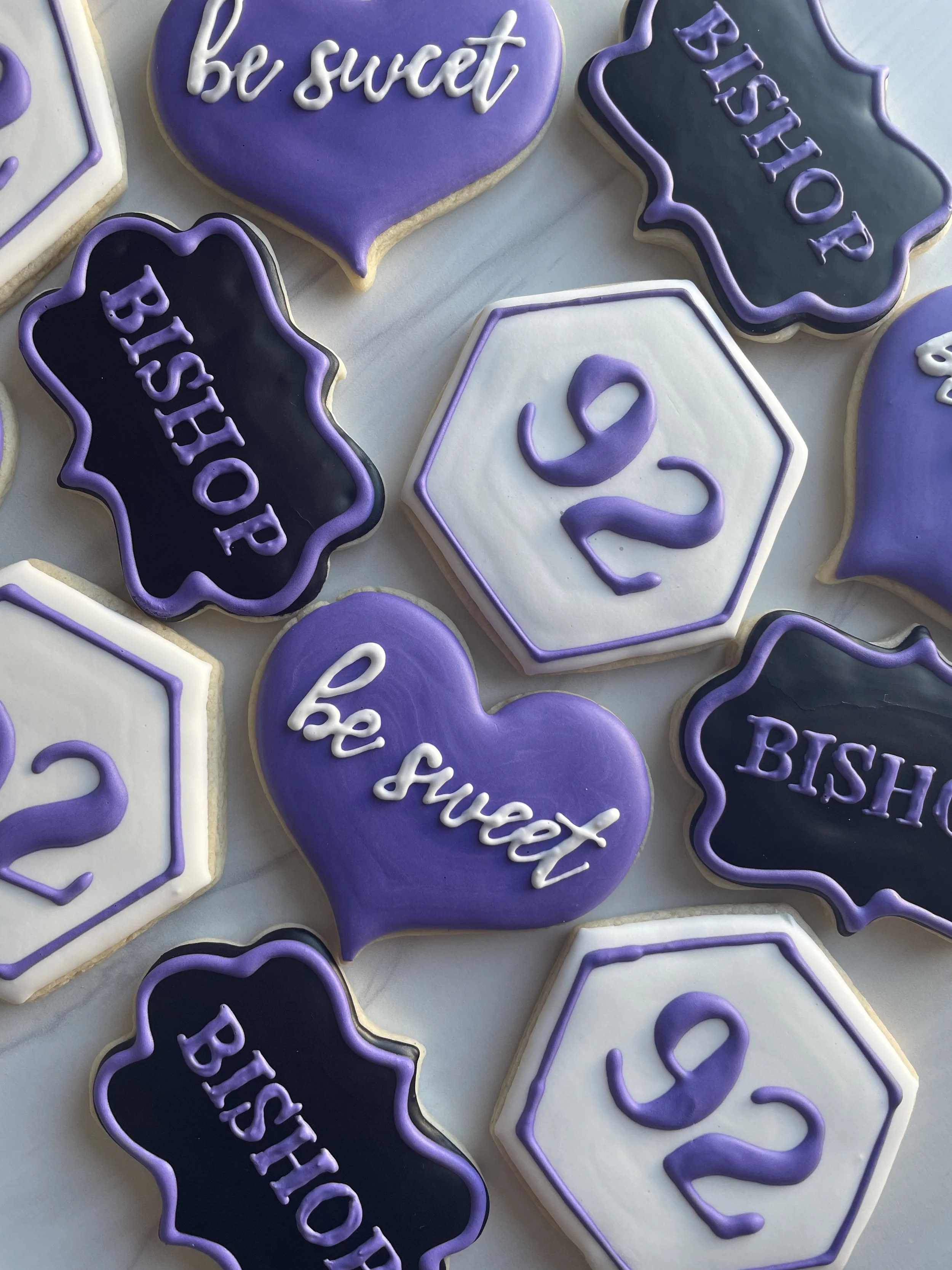 Decorated cookies with messages such as "be sweet," "BISHOP," and the number "2" in purple, black, and white icing, arranged on a light-colored surface.