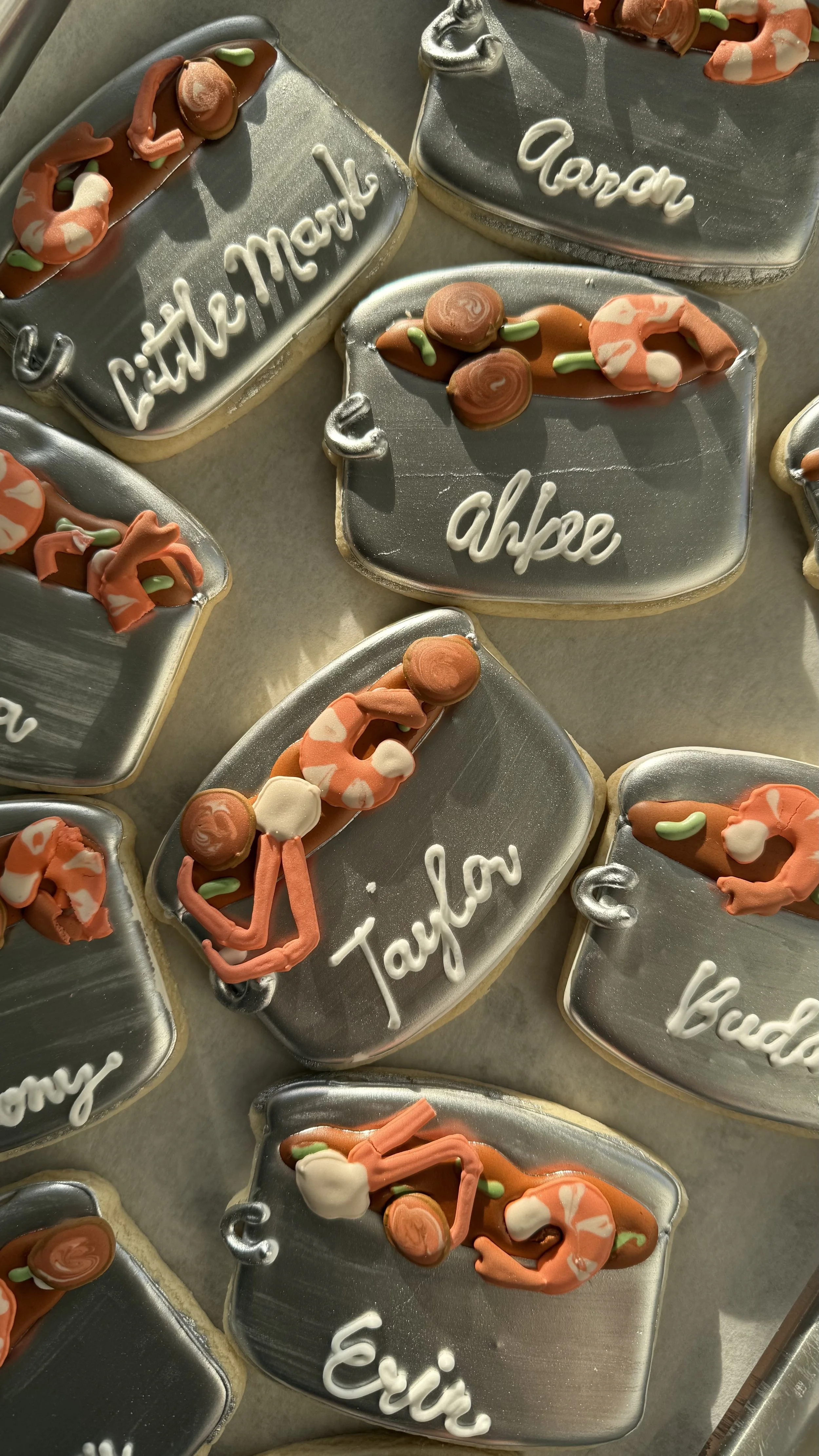 Decorated cookies with silver, orange, white, and green icing, each labeled with names such as 'Taylor', 'Aaron', 'Livi', and 'Eric' on a silver surface.