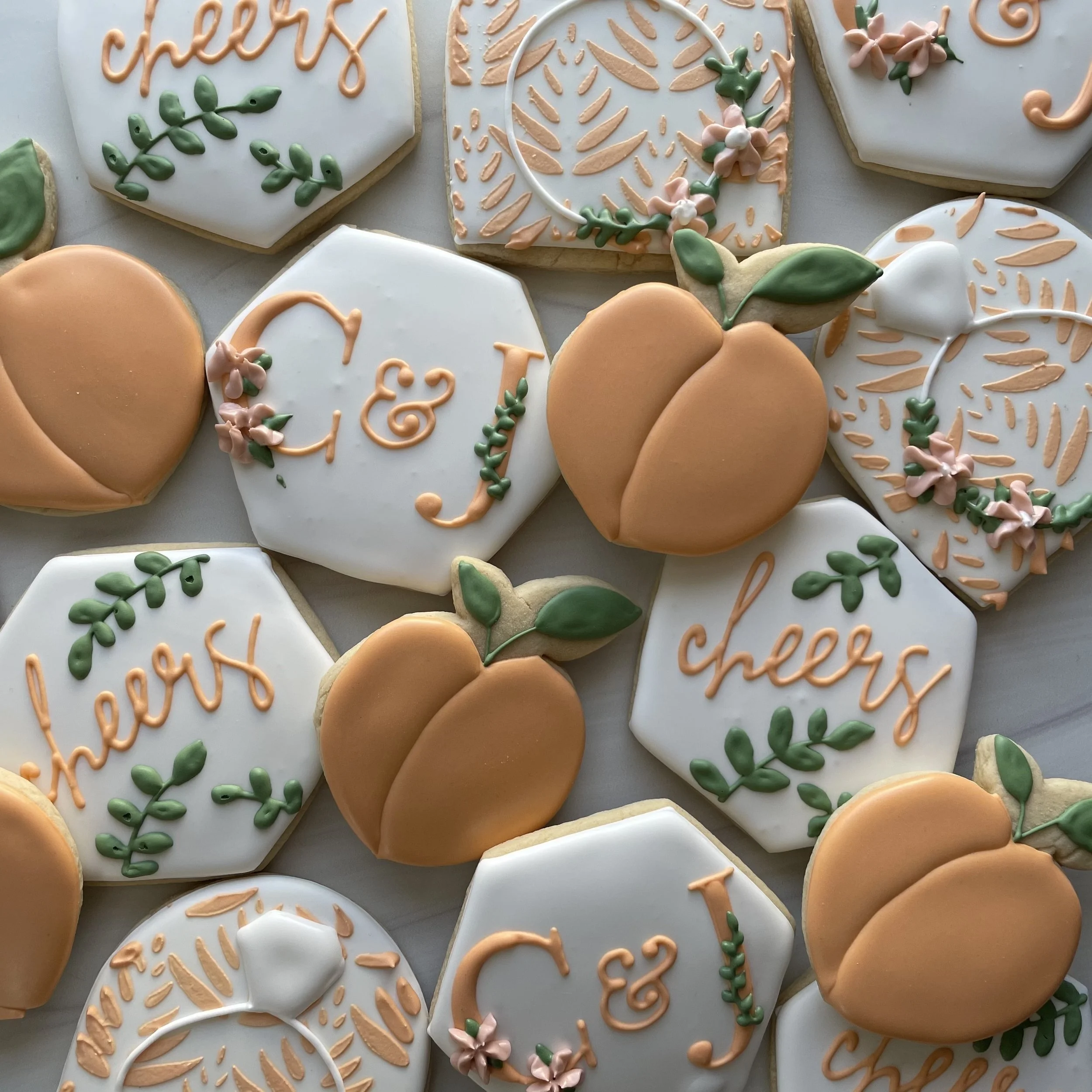 Assorted decorated cookies with flowers, leaves, and cursive writing in peach, pink, and green icing on white icing backgrounds.