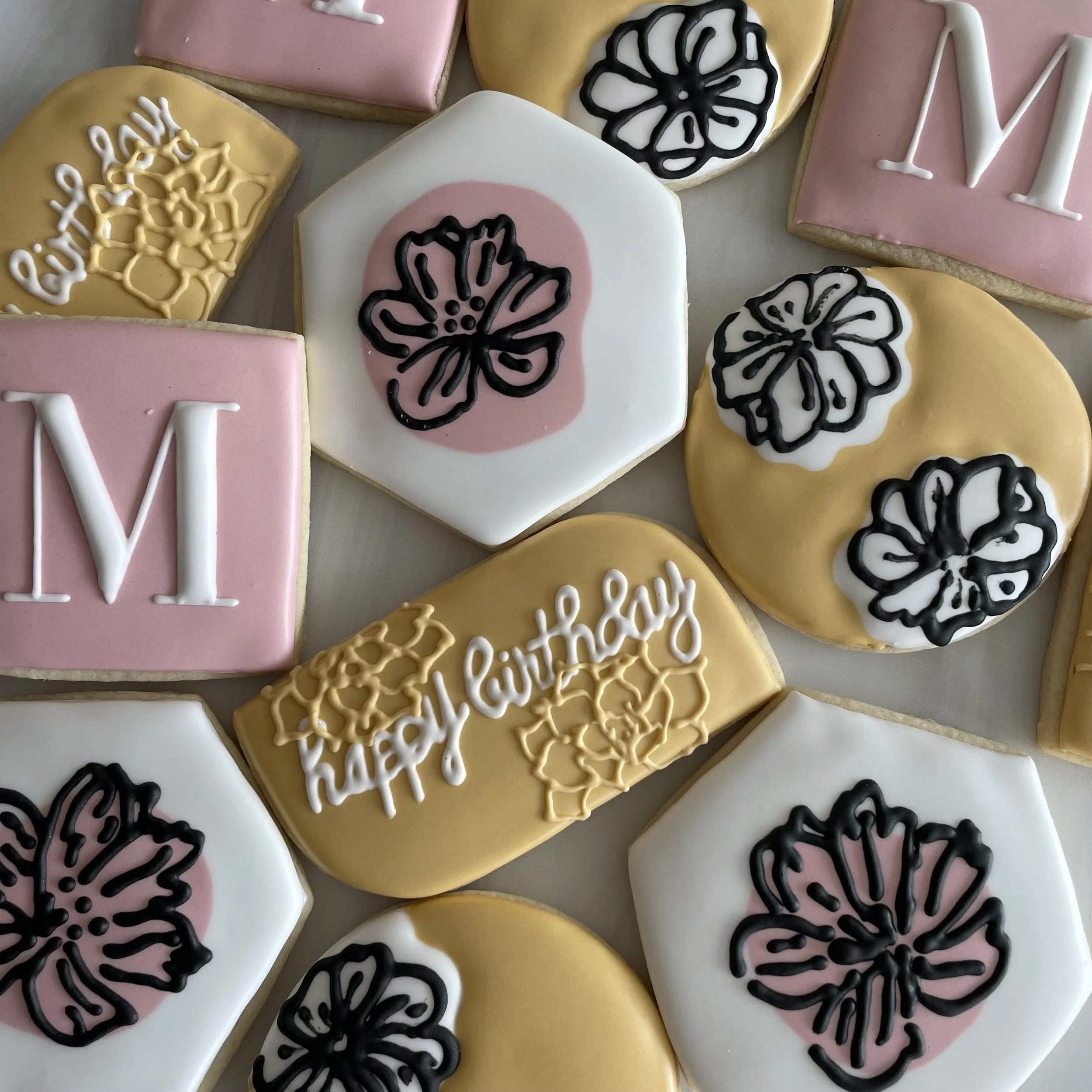 Decorated cookies with pink, white, and yellow icing, black floral designs, and words like 'MOM' and 'birthday' written with white icing.