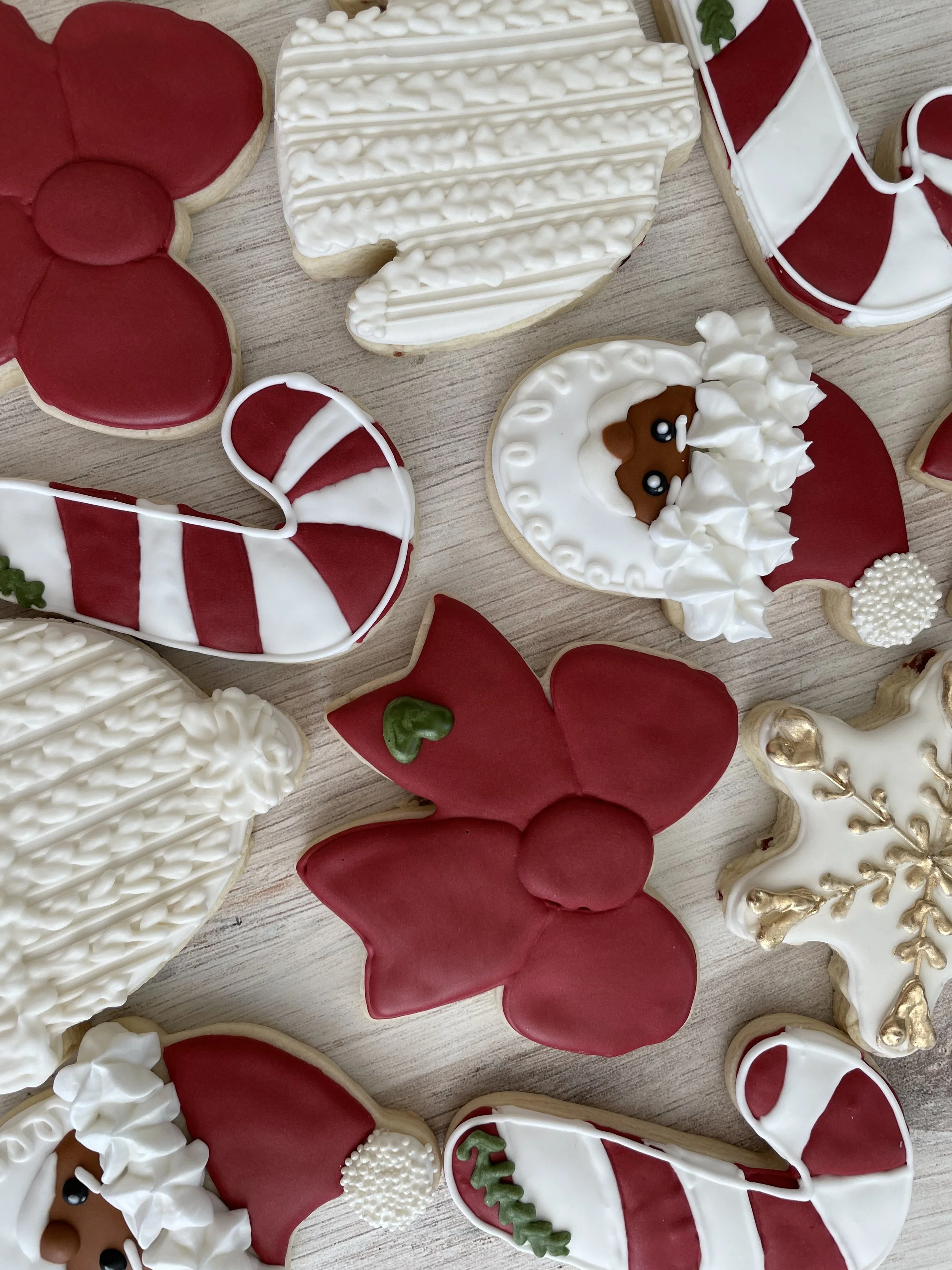 Various decorated Christmas cookies including a red poinsettia flower, a white and red striped candy cane, a heart with red and white icing, a Santa hat, a gingerbread face with white icing, and other festive designs on cookies.