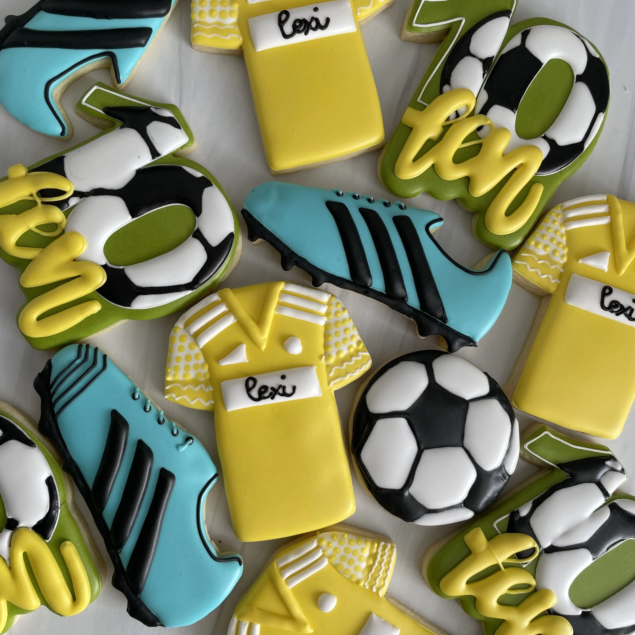 Assorted decorated cookies with sports themes, including soccer balls, soccer jerseys, cleats, and trophies, in yellow, black, white, and blue icing.