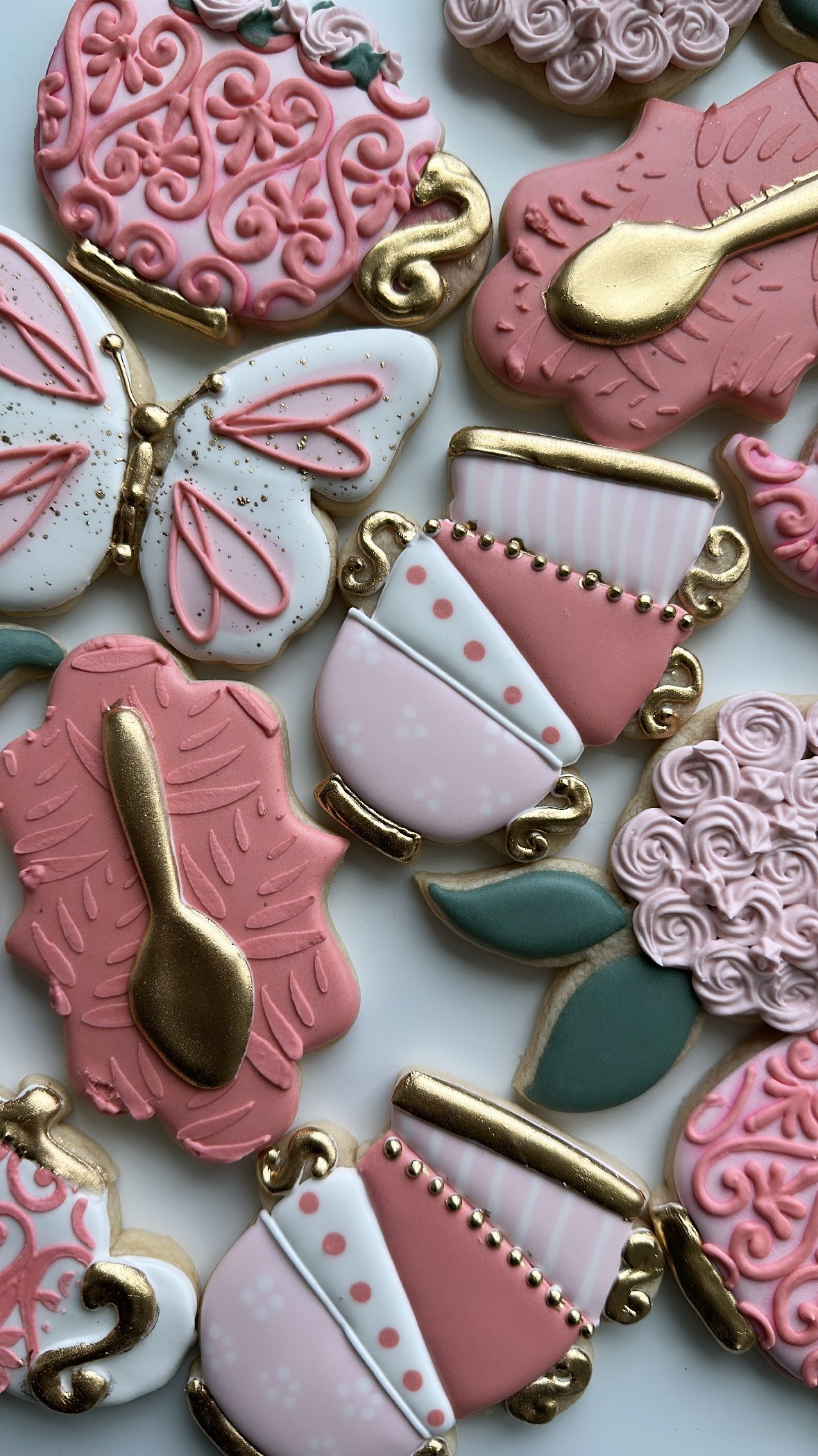 Decorated cookies shaped like teapots, butterflies, and hearts, decorated with pink, white, and gold icing and embellishments.