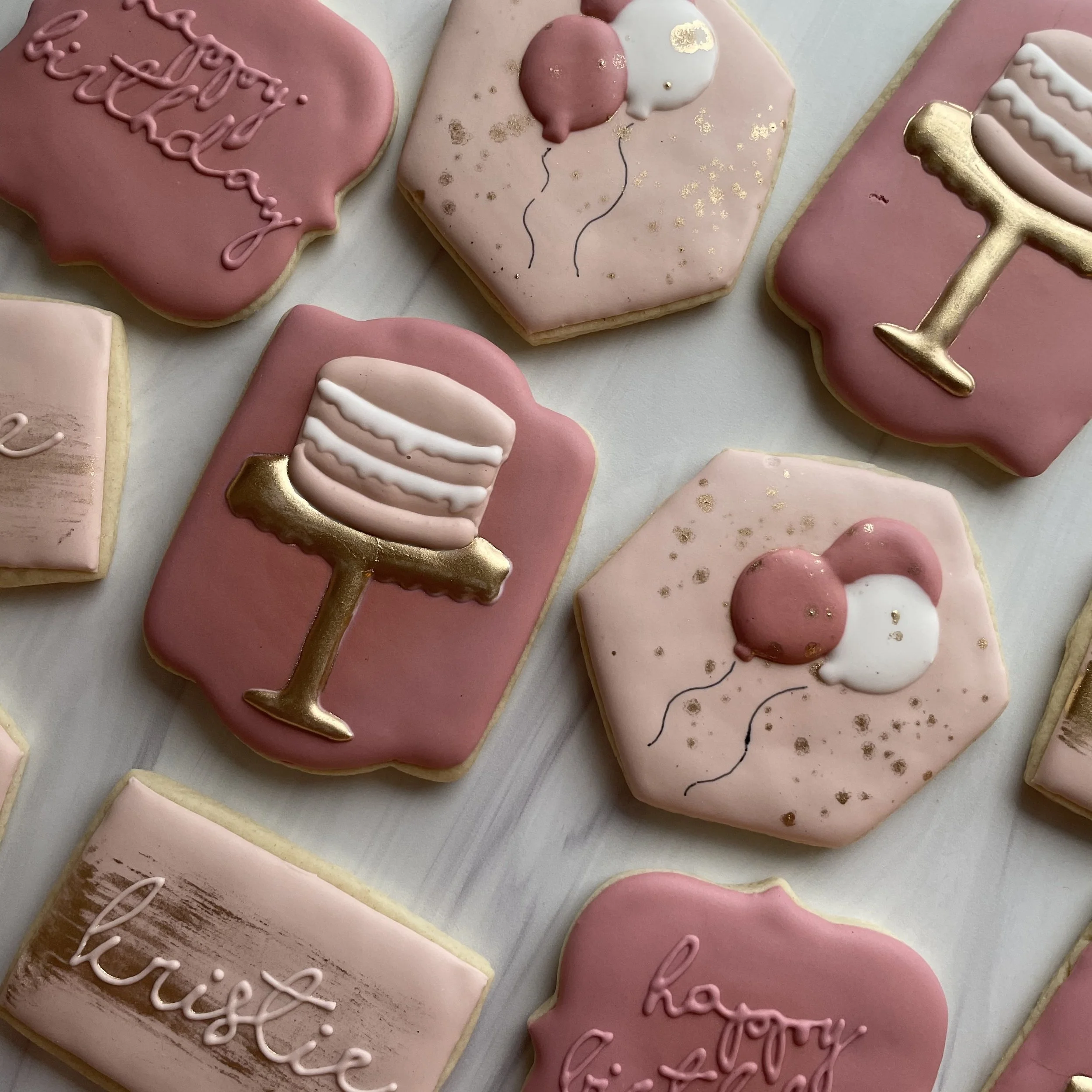Decorated cookies in pink, white, and gold with messages like "Happy birthday" and illustrations such as balloons and drink glasses.