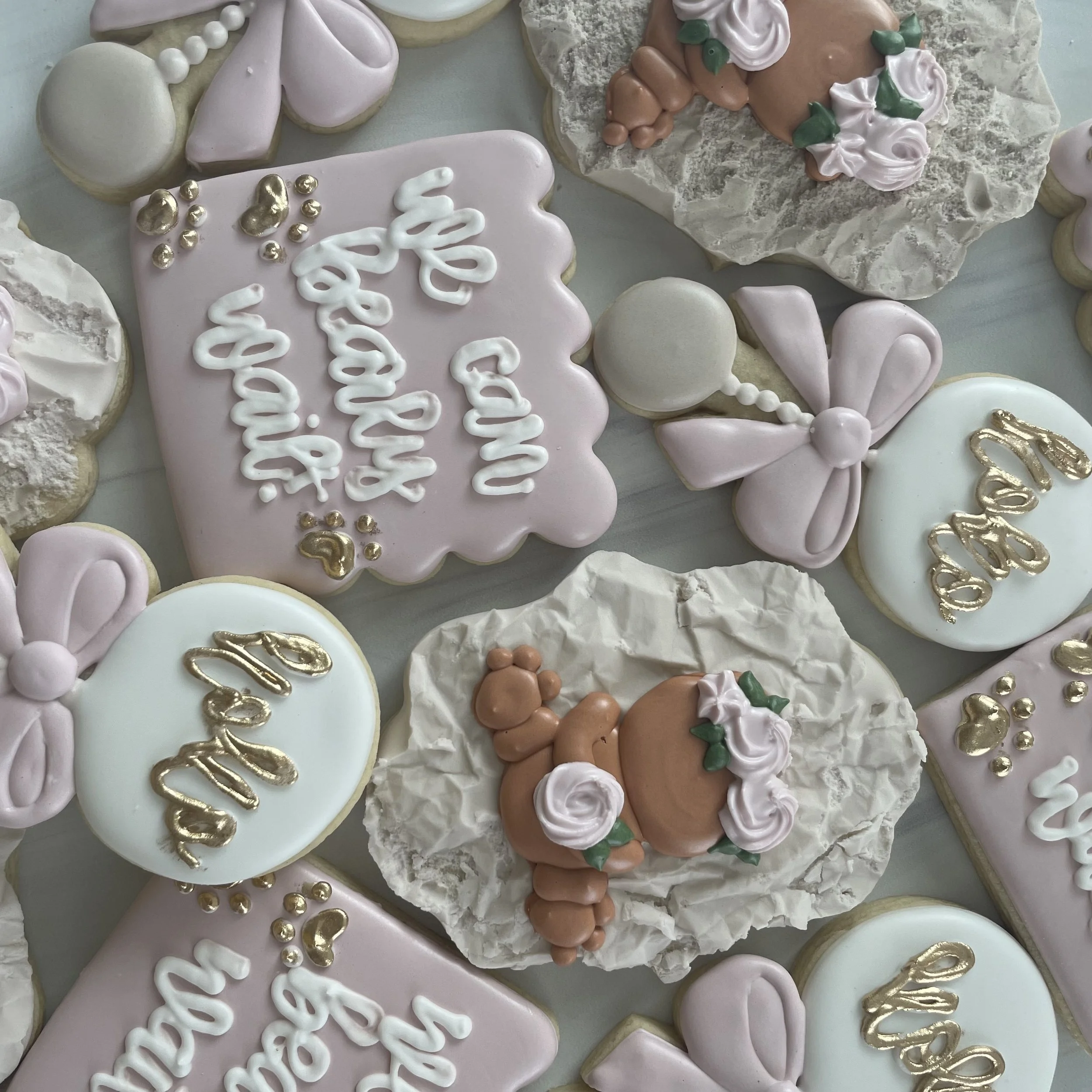 Decorated cookies with pastel pink, white, and brown icing, featuring messages, bows, pearls, and floral designs.