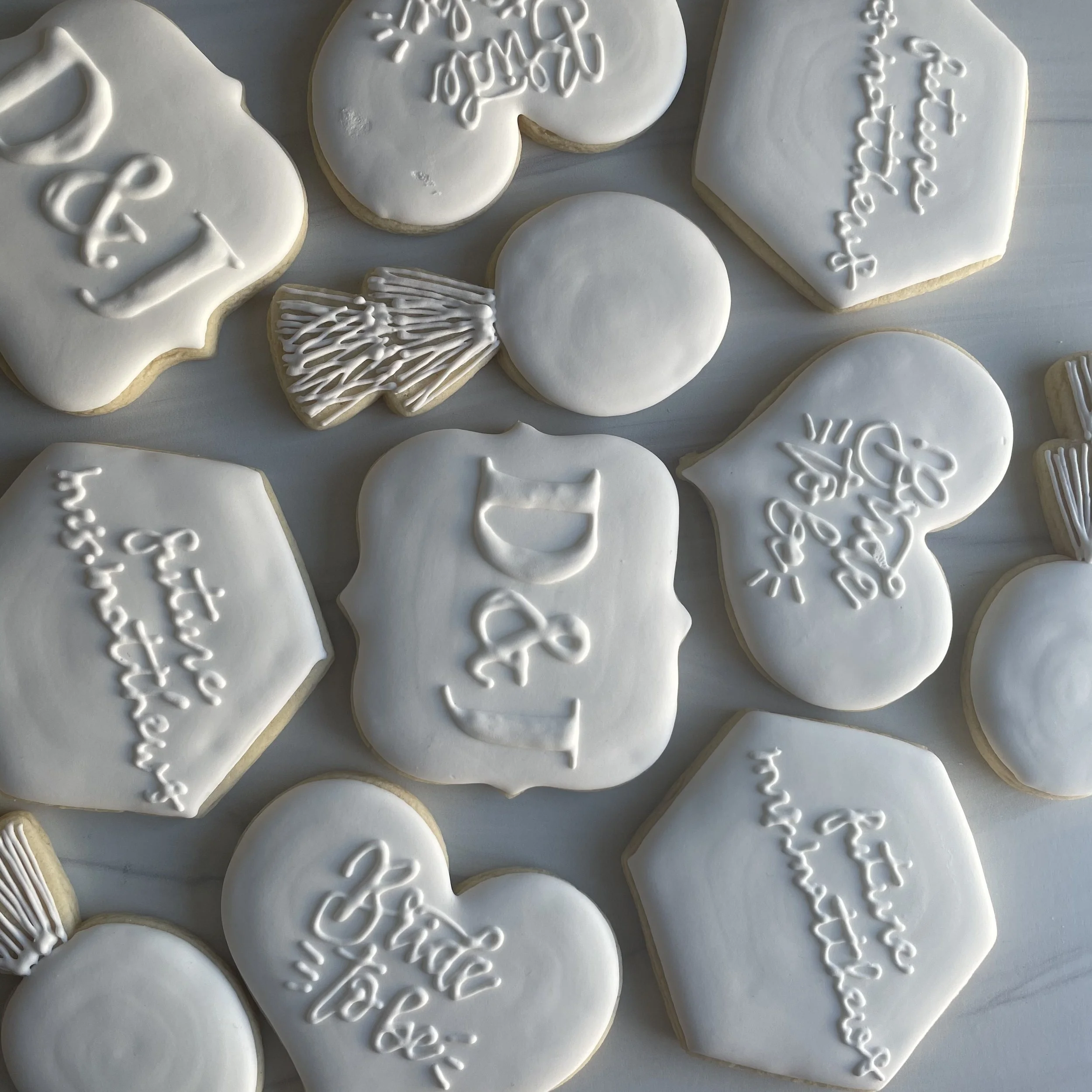 Several white cookies with festive winter and holiday designs, including snowflakes, hearts, and holiday-themed words, decorated with white icing.