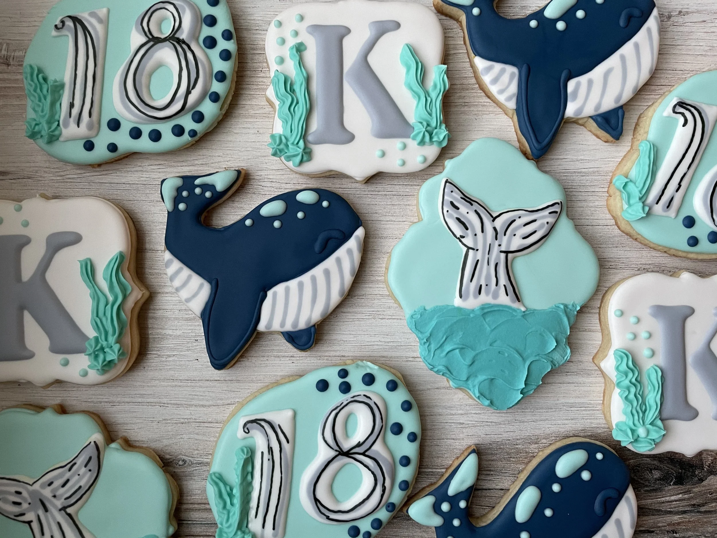 A variety of decorated cookies with ocean and mermaid theme, featuring whales, mermaid tails, and bubbles, in shades of blue, white, and teal, arranged on a wooden surface.