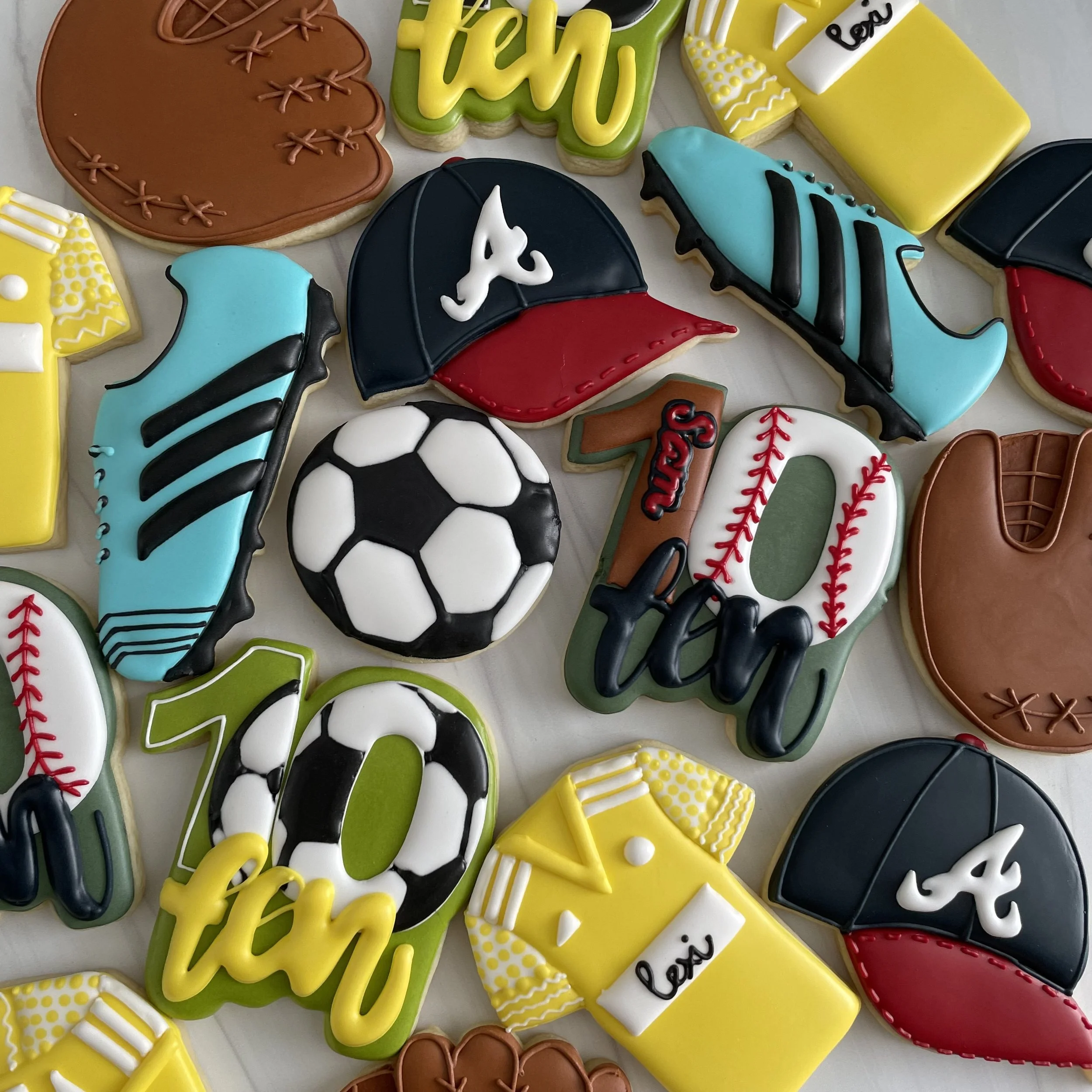 Colorful decorated sugar cookies with sports themes, including baseball gloves, baseballs, footballs, cleats, and words like "play" and "love" in bright yellow, blue, black, red, and brown icing.
