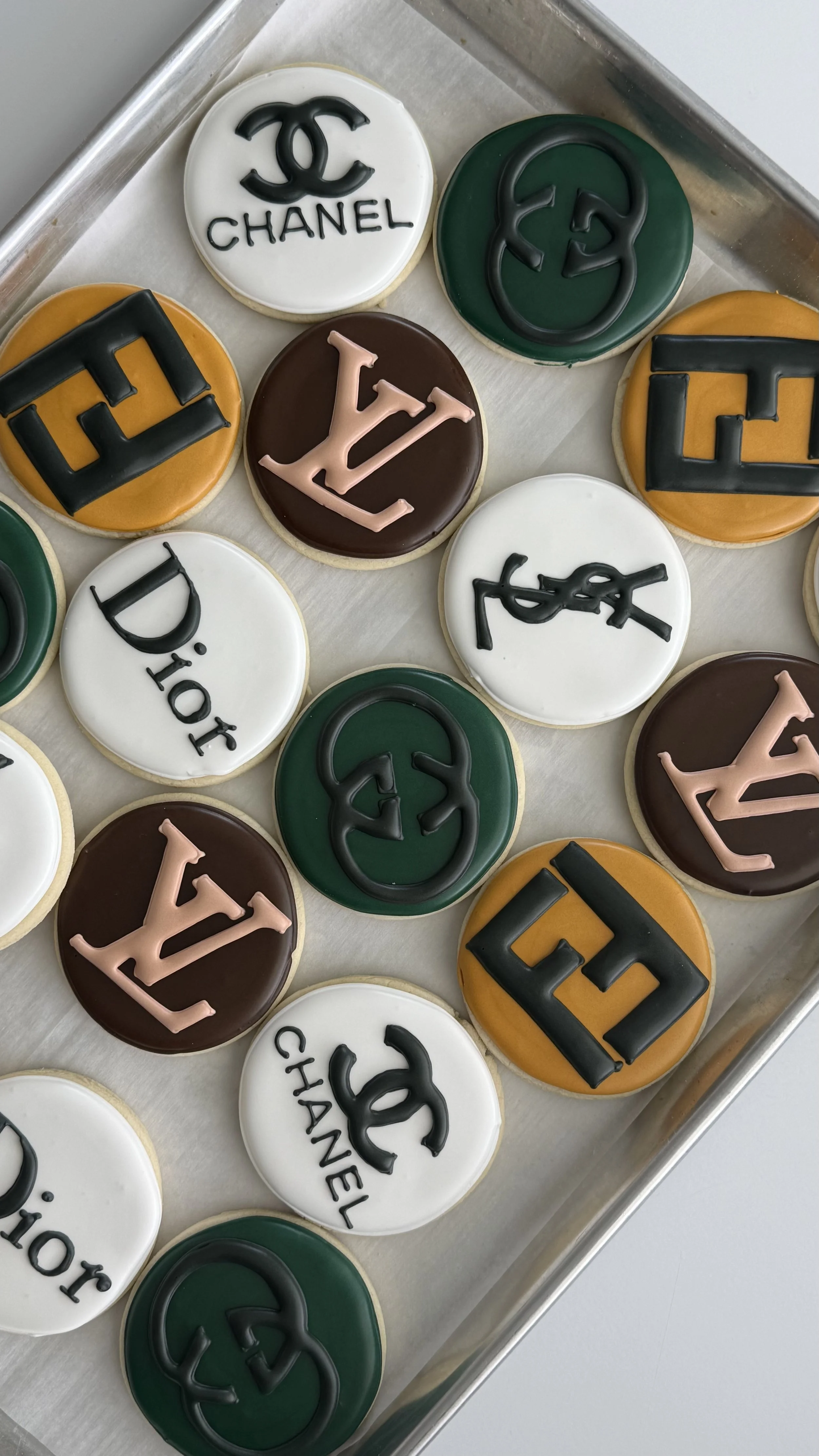 Cookies decorated with logos of Chanel, Gucci, Louis Vuitton, Dior, and Chanel, arranged on a baking sheet.
