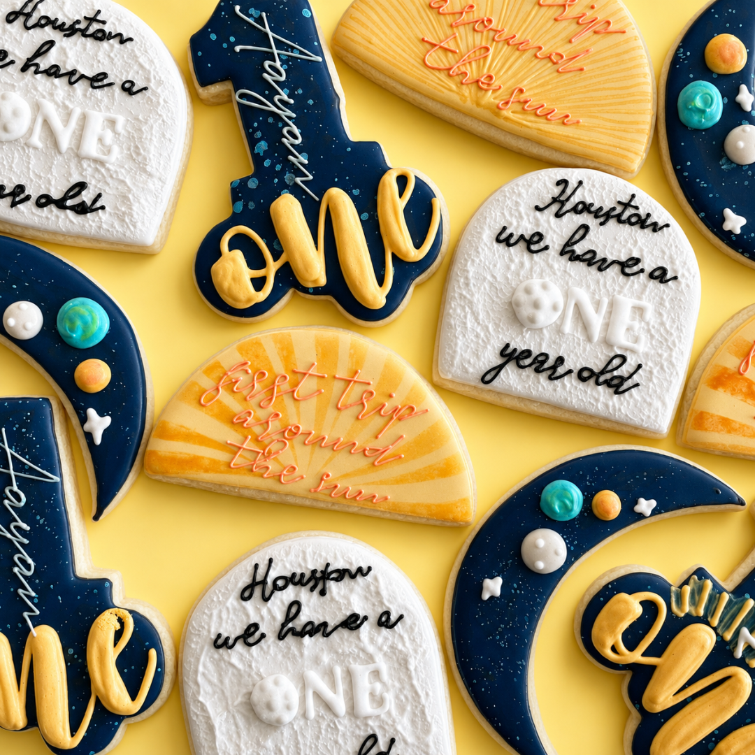 Decorative cookies celebrating a first trip around the sun, with messages like "Houston we have a ONE" and "first trip around the sun," on a yellow background.