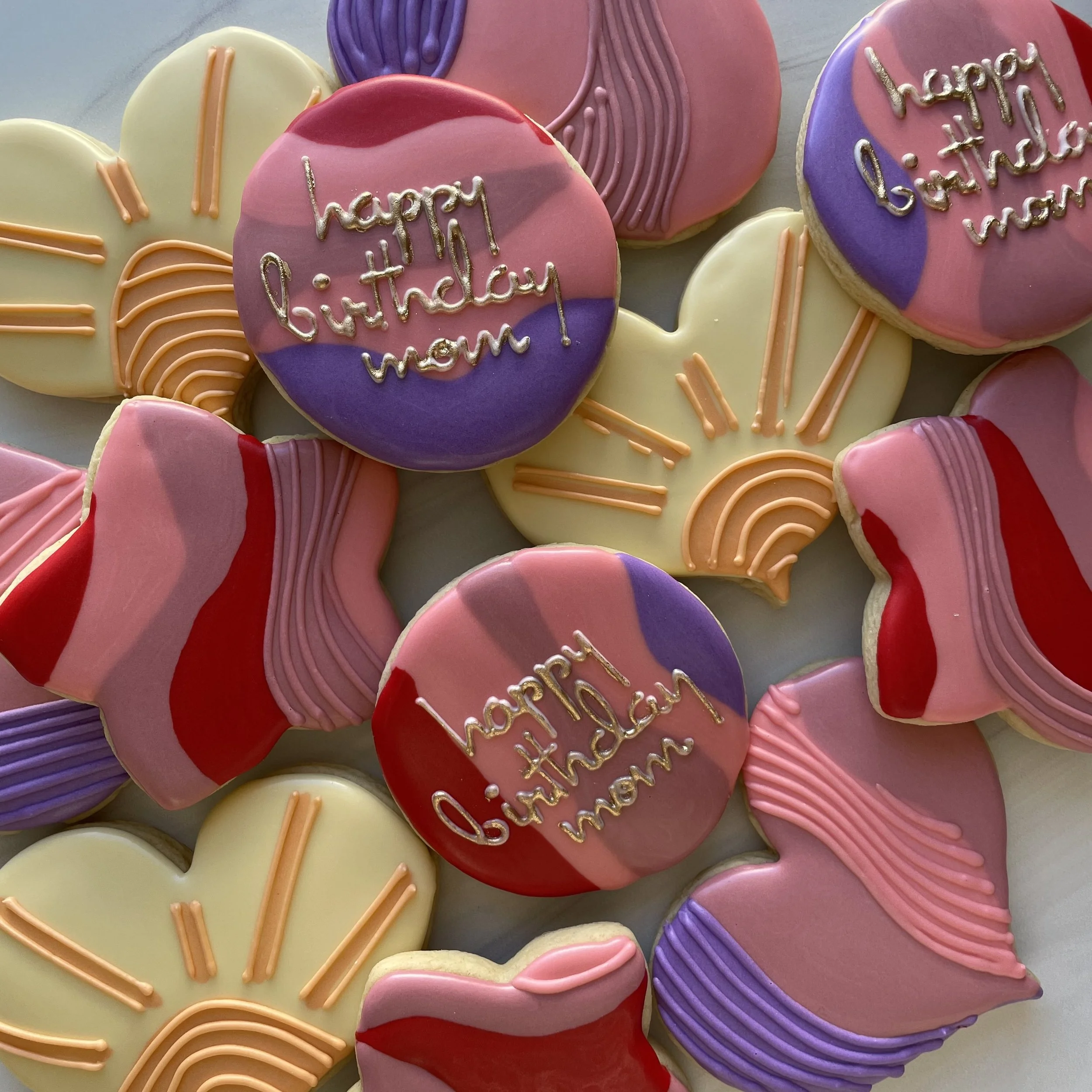 Colorful Heart-shaped cookies with pink, purple, red, and white icing, decorated with various piped lines and the words 'happy birthday mom' written on some of them.