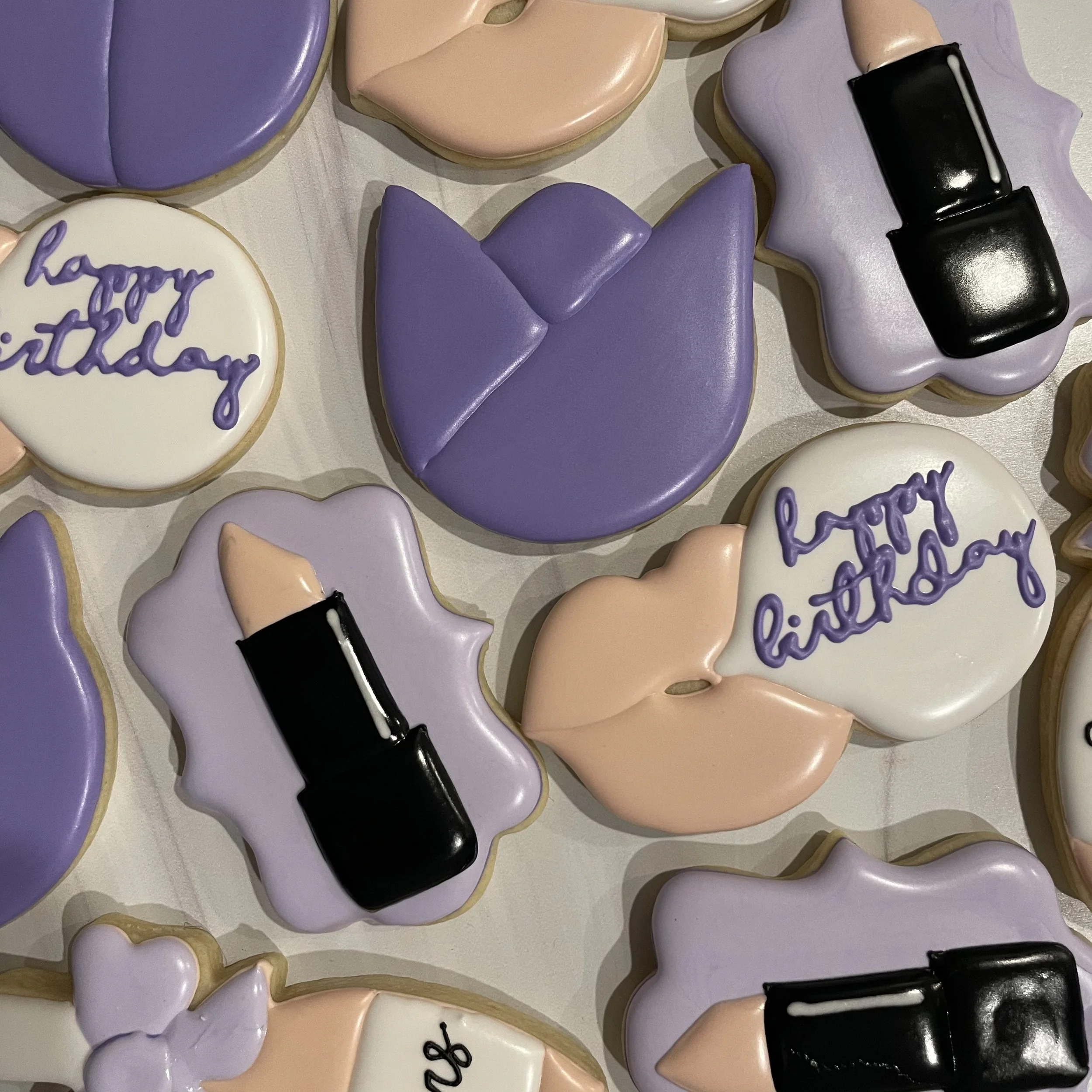 Decorated cookies in pastel purple, pink, and white colors for a birthday celebration. Some cookies have messages written in purple icing, and others are shaped like hearts, bows, and lipstick.