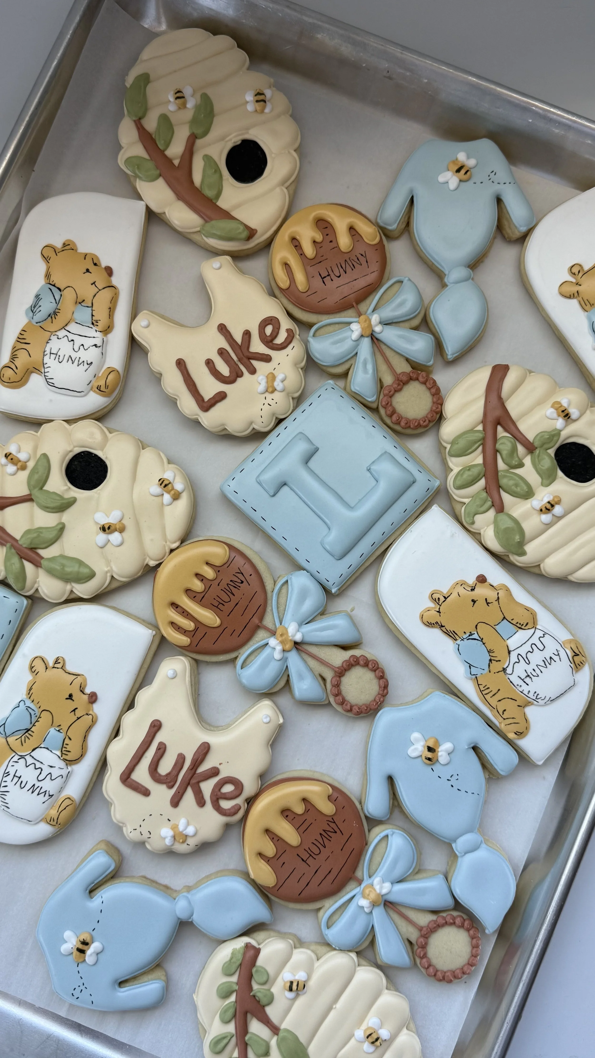 Decorated cookies with honey and bear themes, including a bear holding a jar labeled "Hunny," honey dipper, bees, a blue sweater, and the letter "T" on a blue background.